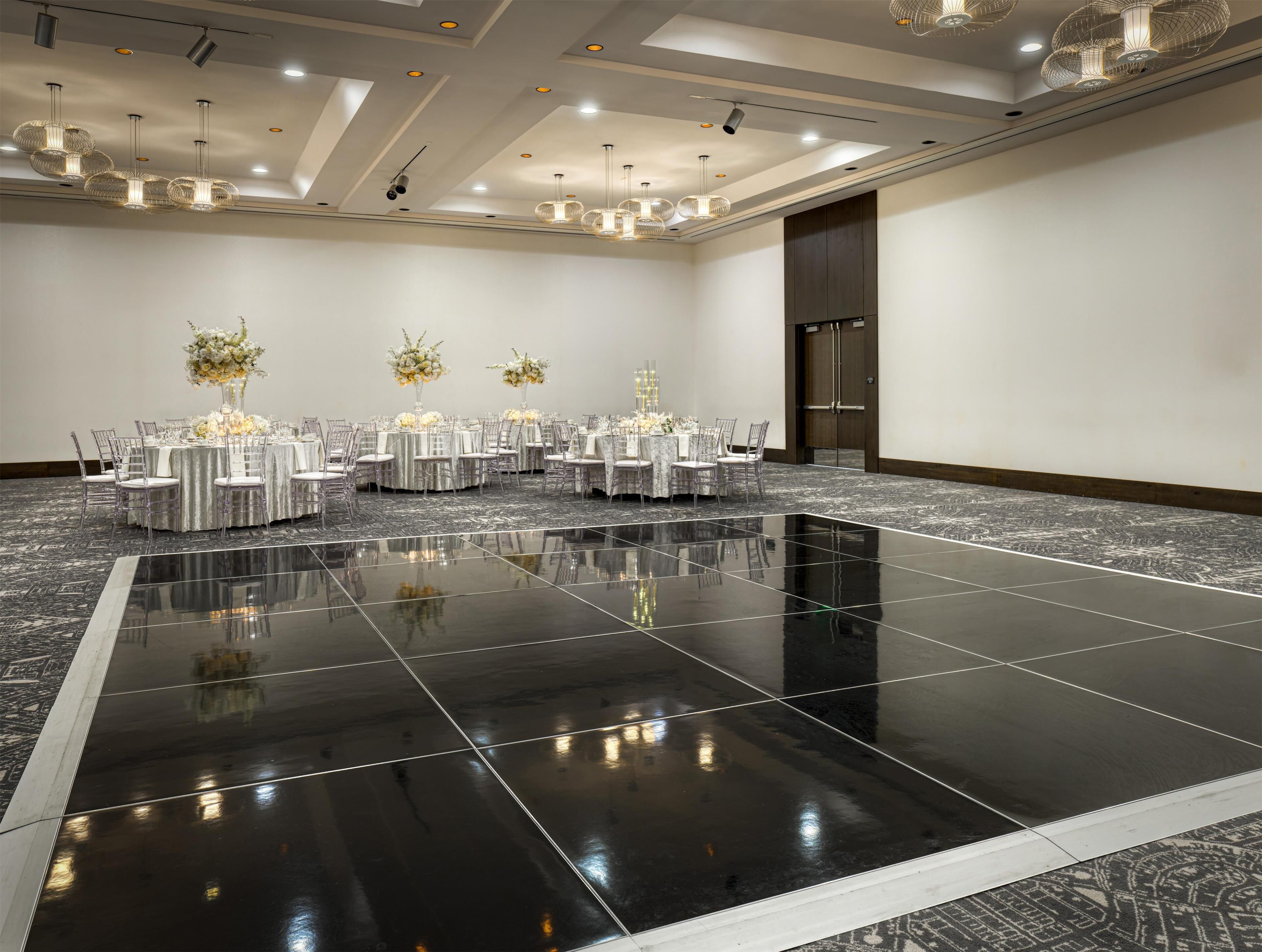 Un amplio salón de banquetes, bien iluminado, con varias mesas redondas, sillas transparentes y elegantes centros de mesa florales.