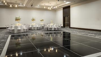 Un amplio salón de banquetes, bien iluminado, con varias mesas redondas, sillas transparentes y elegantes centros de mesa florales.