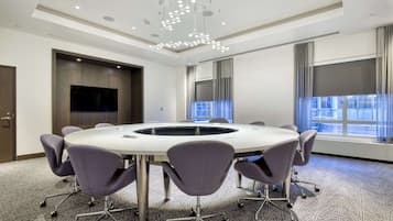 Uma sala de conferências com uma mesa redonda, seis cadeiras, uma TV de tela plana e uma janela grande com cortinas.