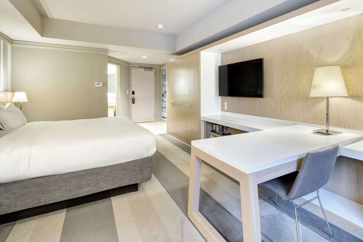 Una habitació d'hotel moderna amb un llit, un escriptori, una cadira, un televisor i una làmpada.