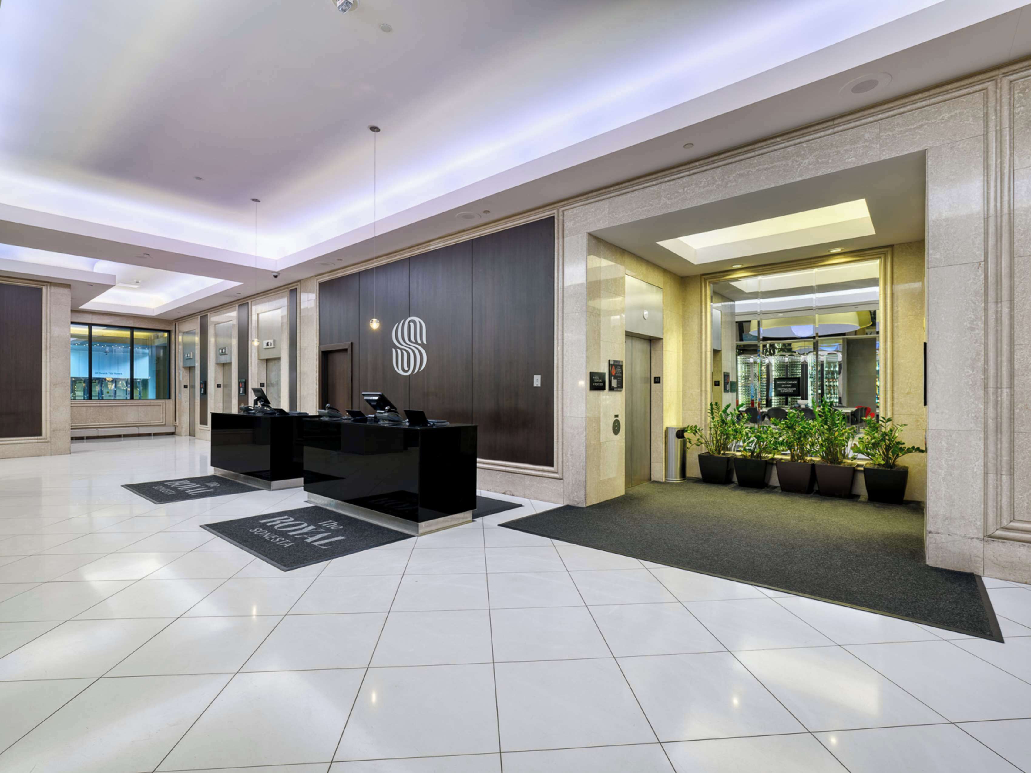 A modern hotel lobby with a reception desk, elevators, and potted plants.