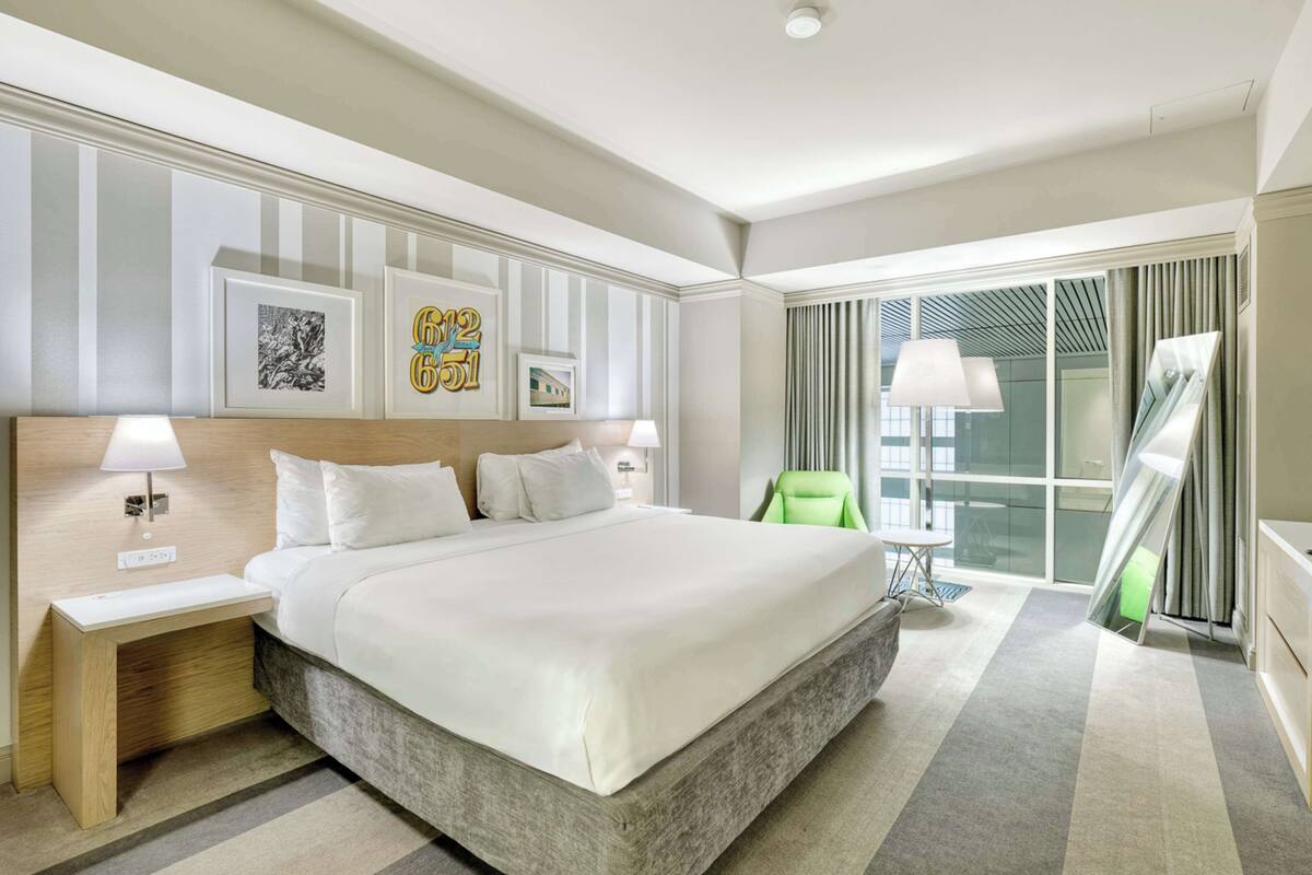 A hotel room with a large bed, a bedside table, a green chair, and a large window with curtains.