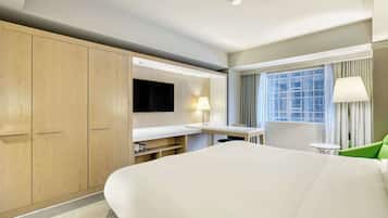 Quarto de hotel com uma cama grande, uma mesa com cadeira, TV fixada na parede e uma janela com cortinas.