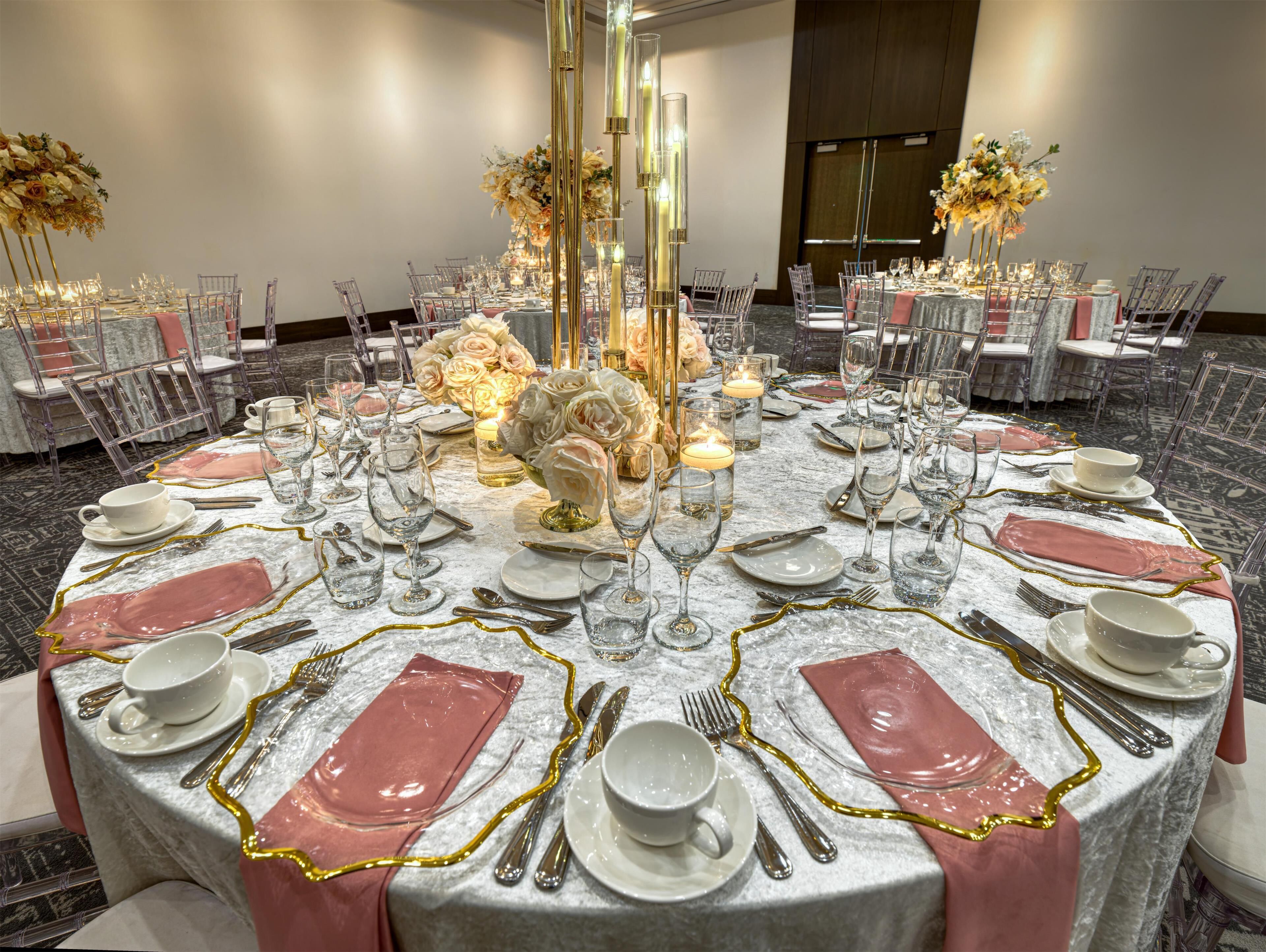 Una mesa redonda dispuesta para un evento formal, con copas transparentes, platos blancos y servilletas rosas.