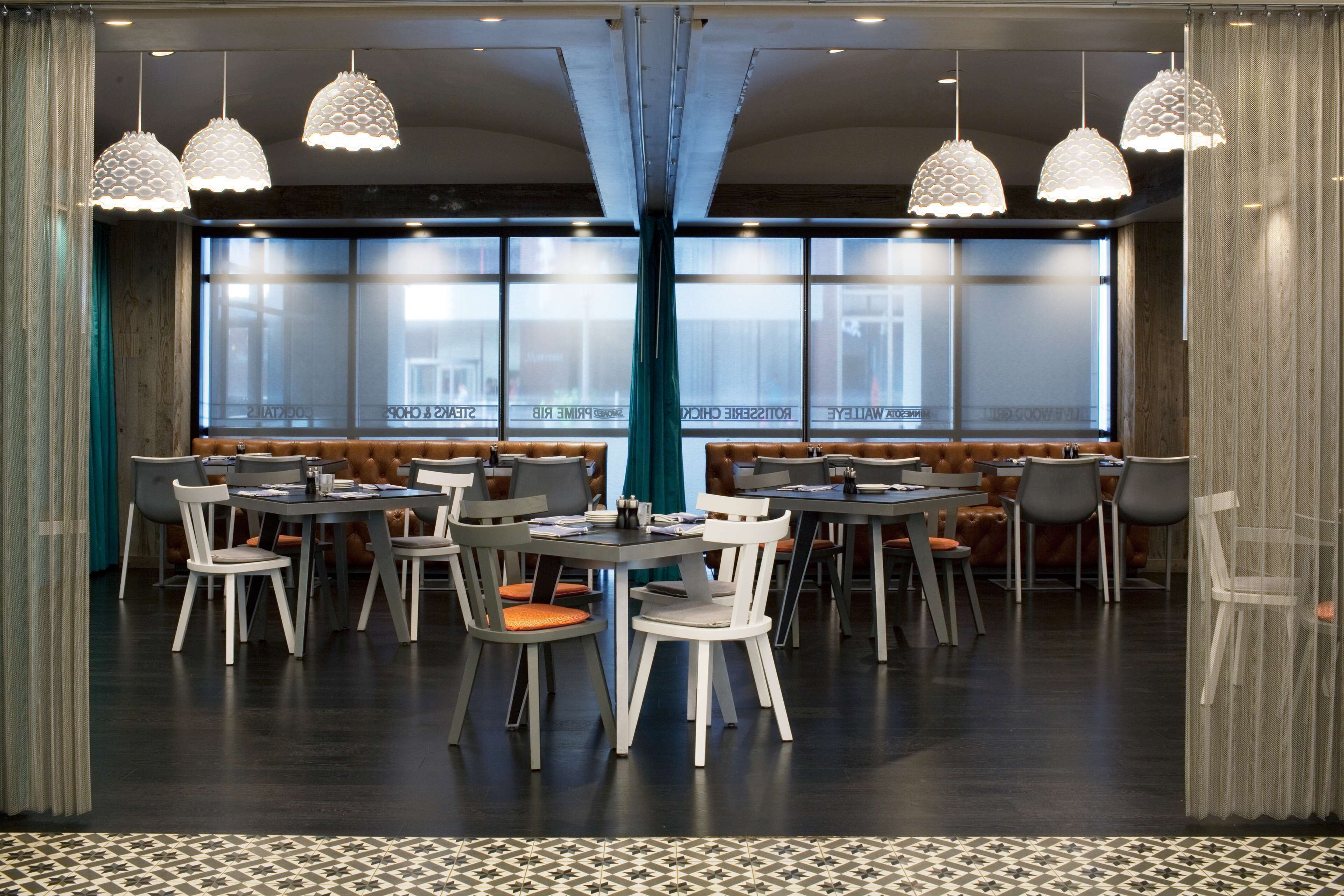 Een moderne eethoek met meerdere tafels en stoelen, grote ramen en decoratieve hanglampen.