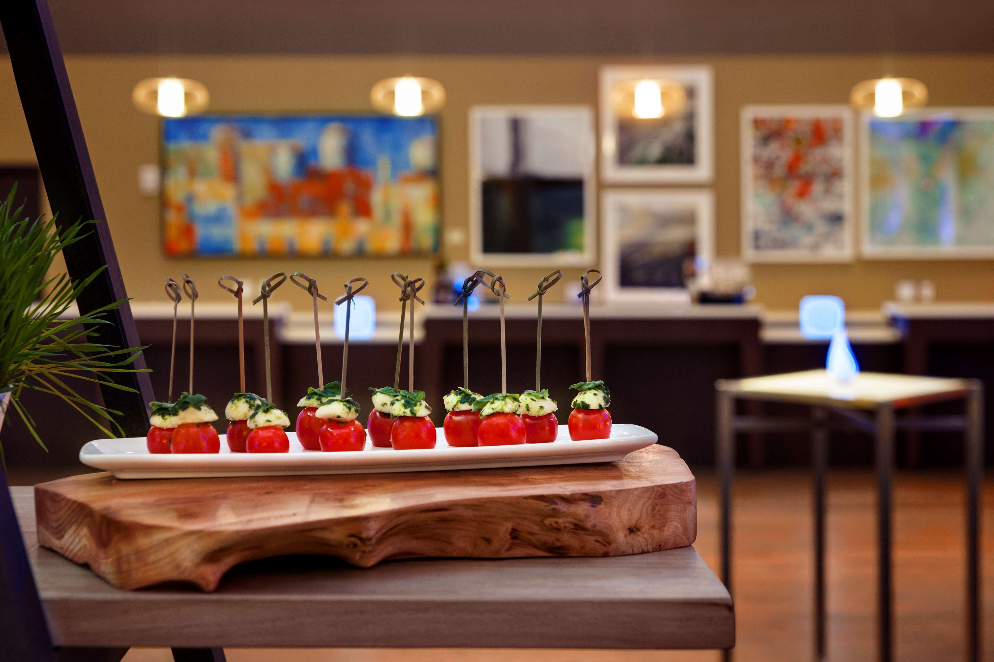 Een houten serveerplank met spiesen kersentomaten, versierd met kruiden, op een tafel in een restaurant met kunstwerken aan de muur.