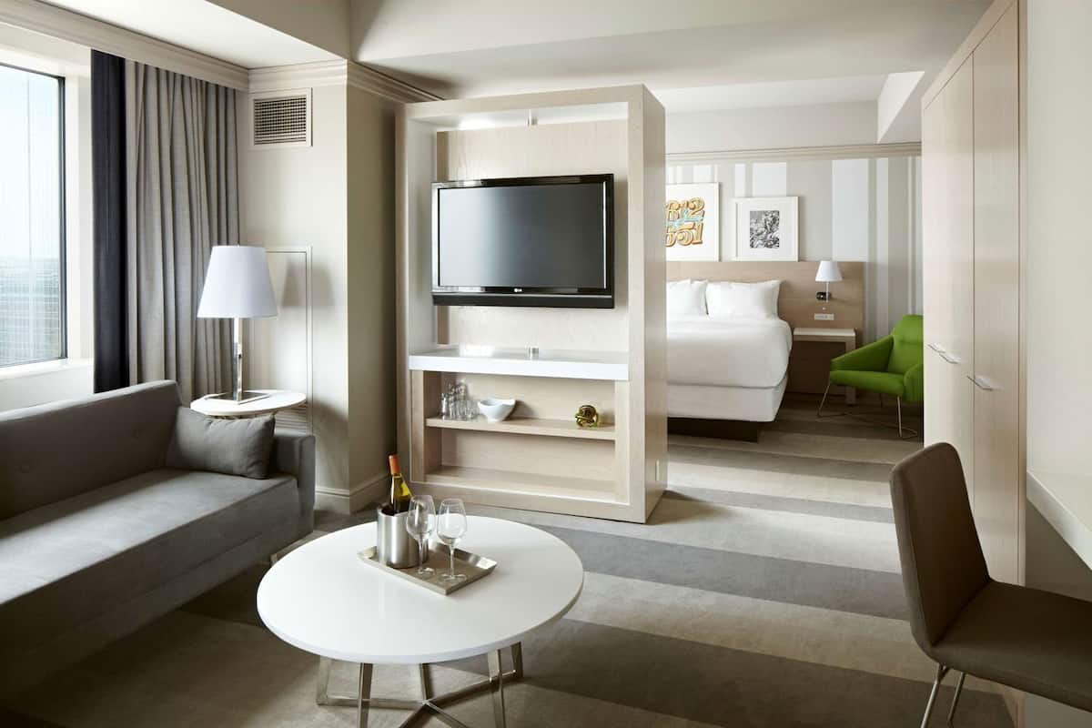 A hotel room with a sofa, a round table with a bottle and glasses, a TV, a bed, and a green chair.