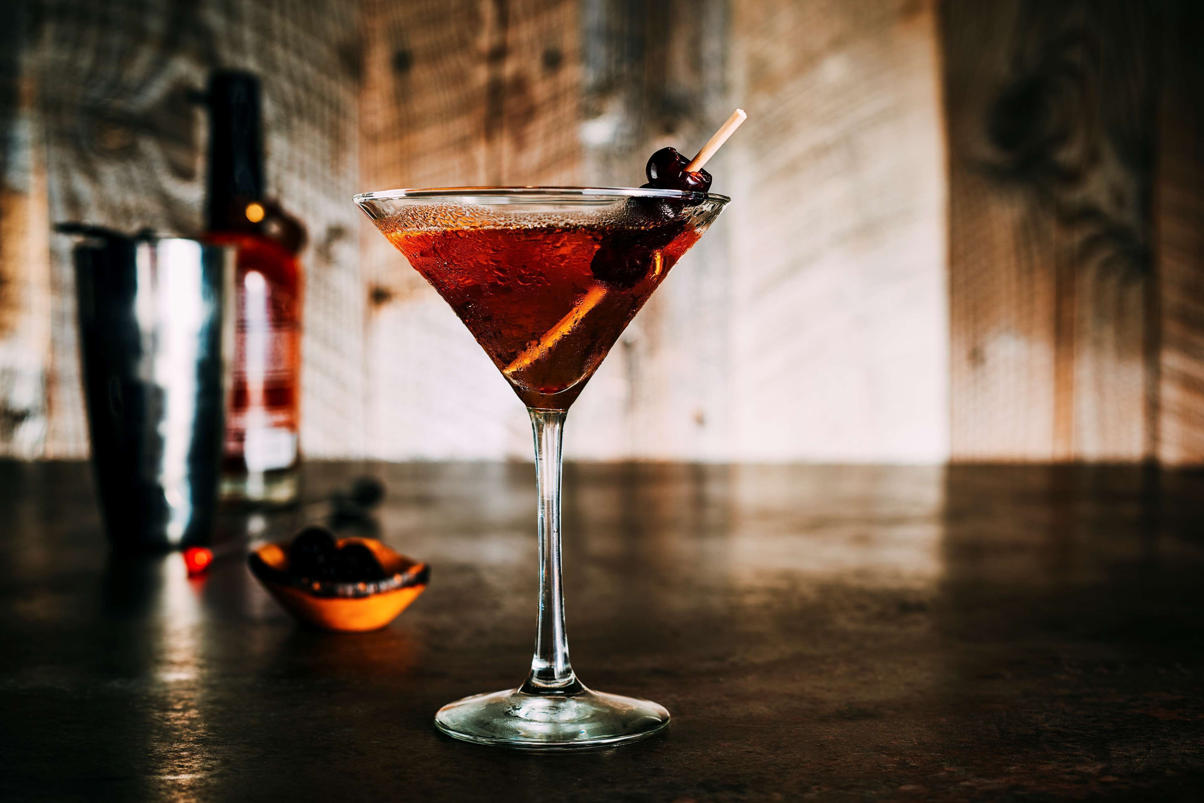 Una copa de martini con una cereza en un palillo, una botella de licor y un cuenco de aceitunas sobre una superficie de madera.
