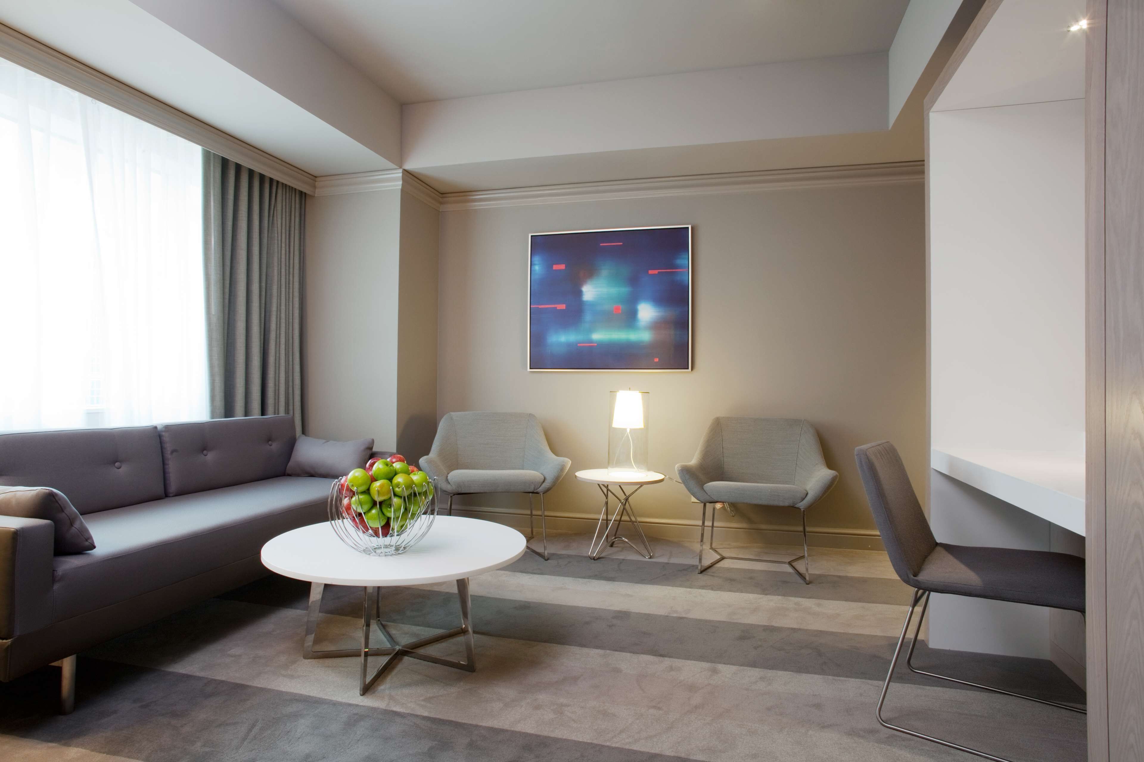 A modern hotel room with a sofa, two armchairs, a small table with a vase of flowers, a lamp, and a painting on the wall.