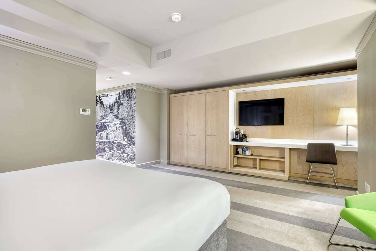 A modern hotel room with a large bed, a desk with a flat-screen TV, and a green chair.