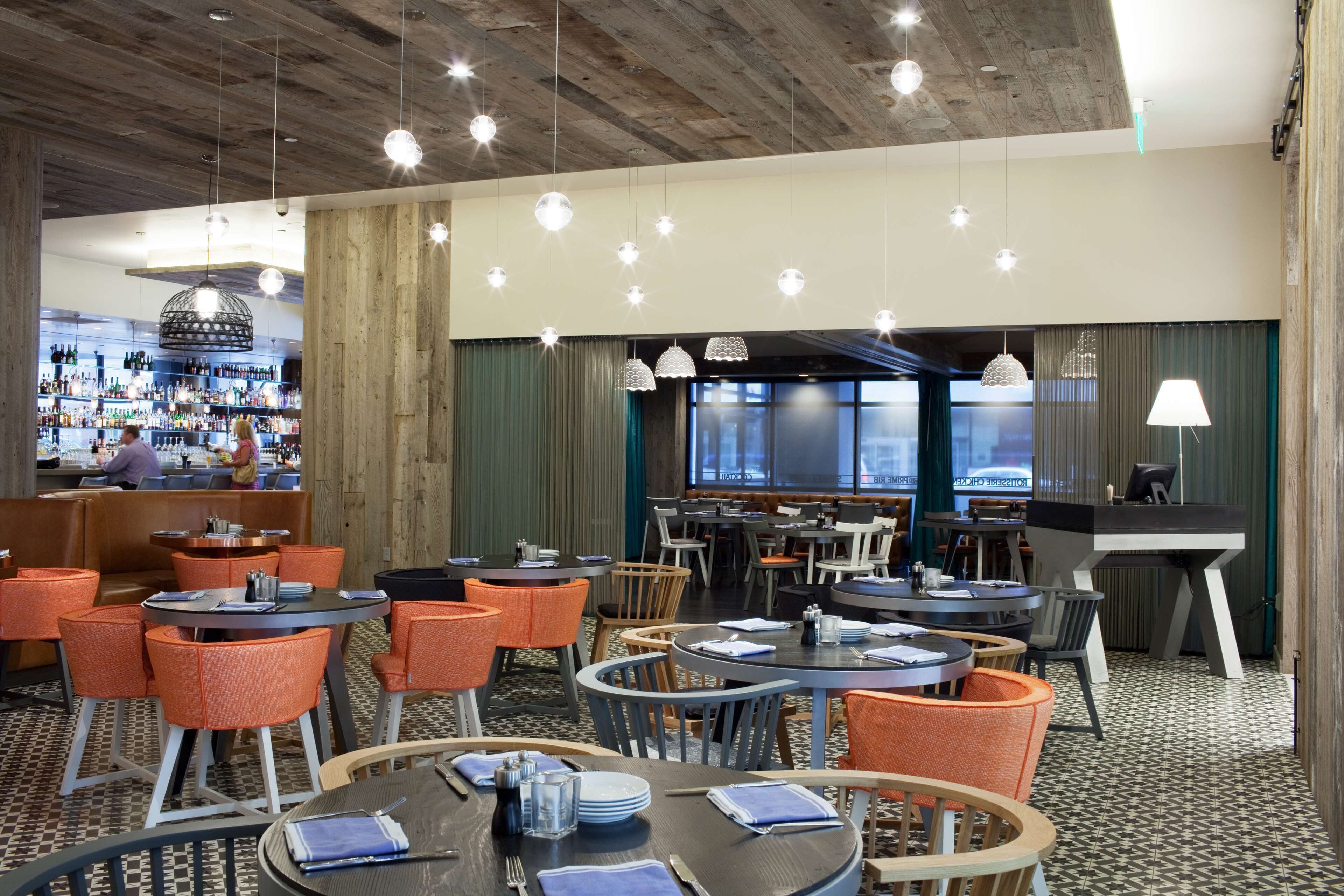 Un vestíbulo de hotel con mesas redondas y sillas naranjas, una zona de bar con estantes llenos de botellas y un ventanal con cortinas.