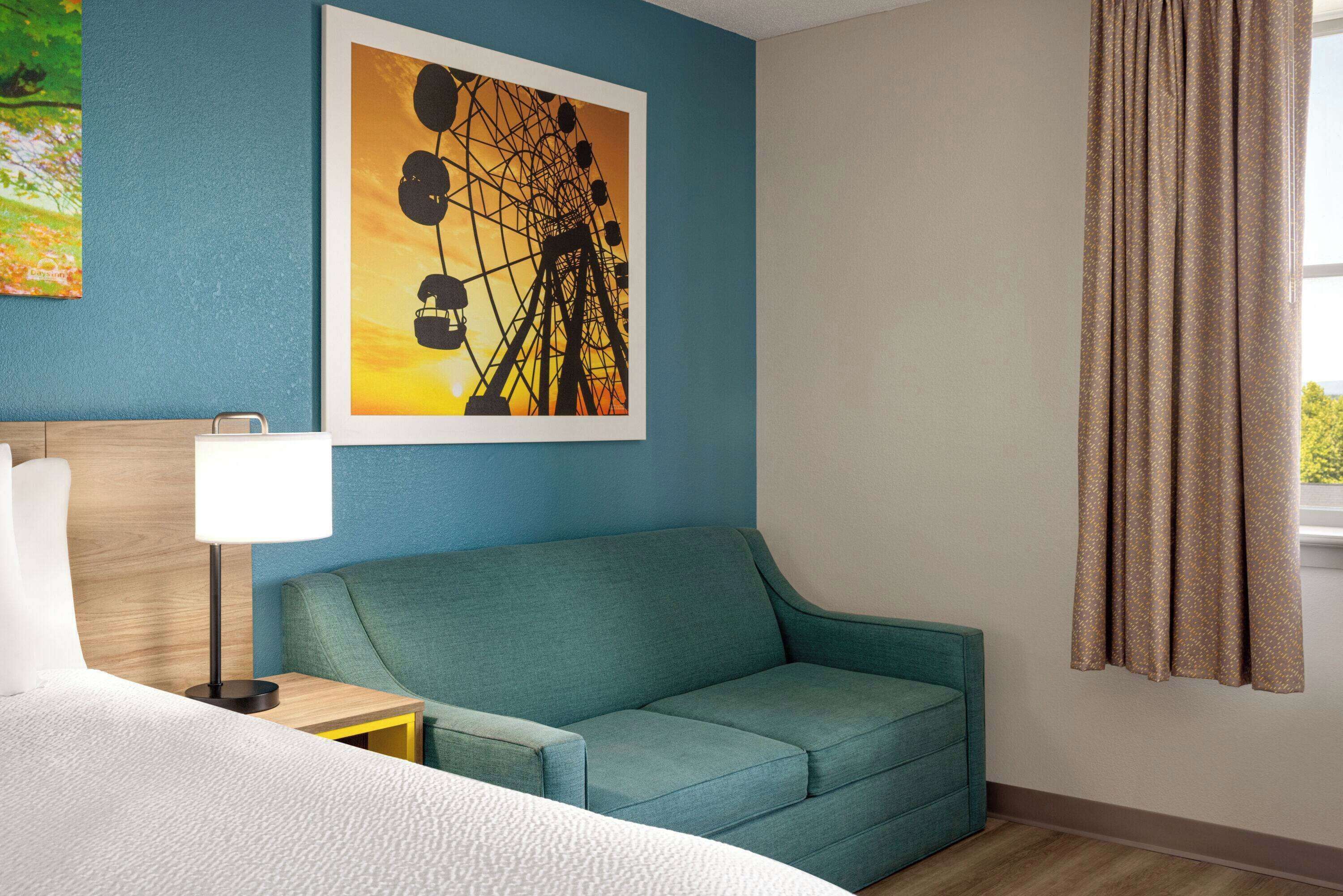 Habitación de hotel con cama, sofá color verde azulado, cuadro enmarcado de una noria, mesita de noche con lámpara y ventana con cortinas.
