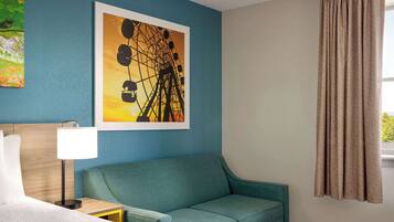 Habitación de hotel con cama, sofá color verde azulado, cuadro enmarcado de una noria, mesita de noche con lámpara y ventana con cortinas.
