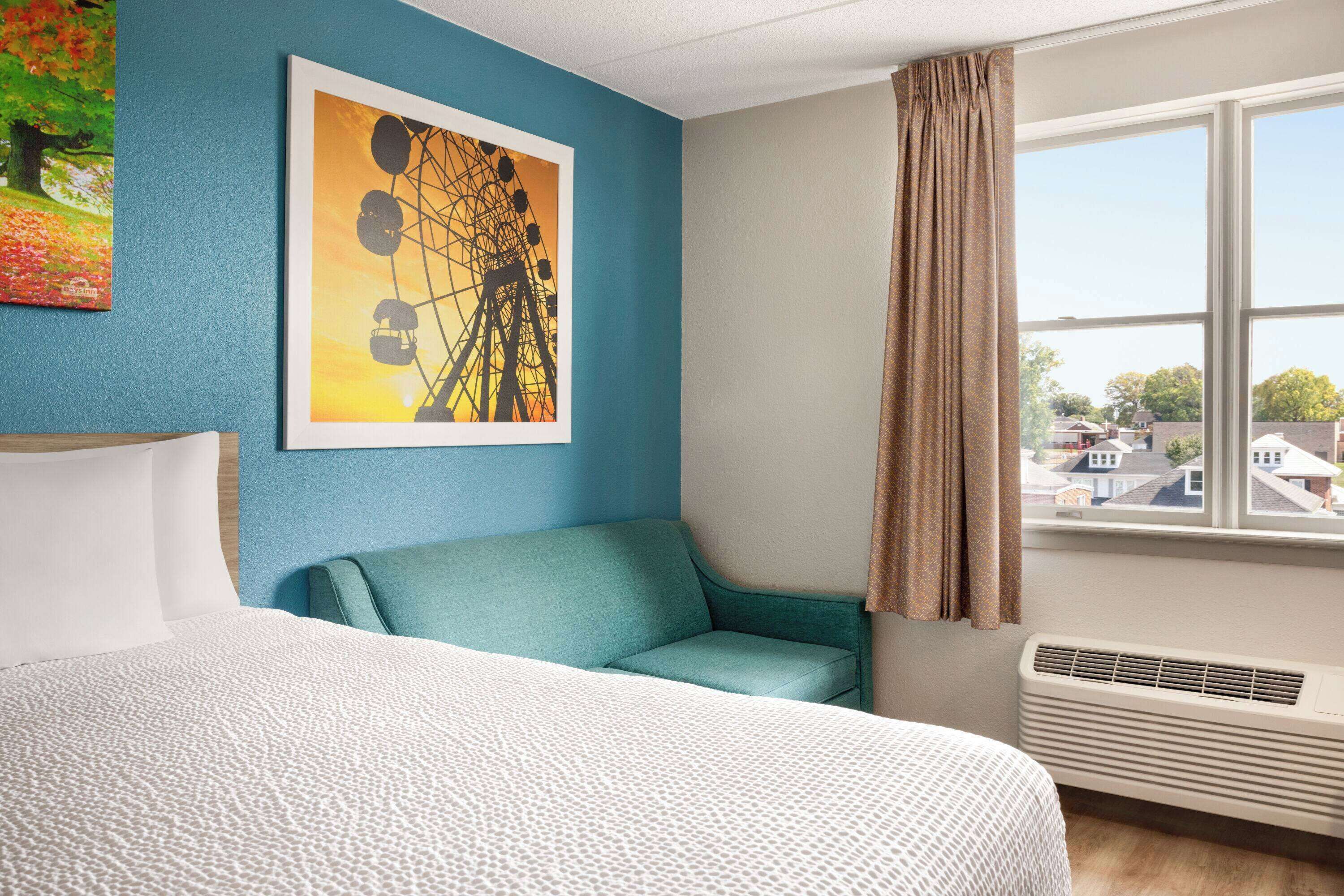 Una habitación de hotel con una cama, un sillón color verde azulado, un ventanal con vista a casas y una pared con un cuadro de noria.