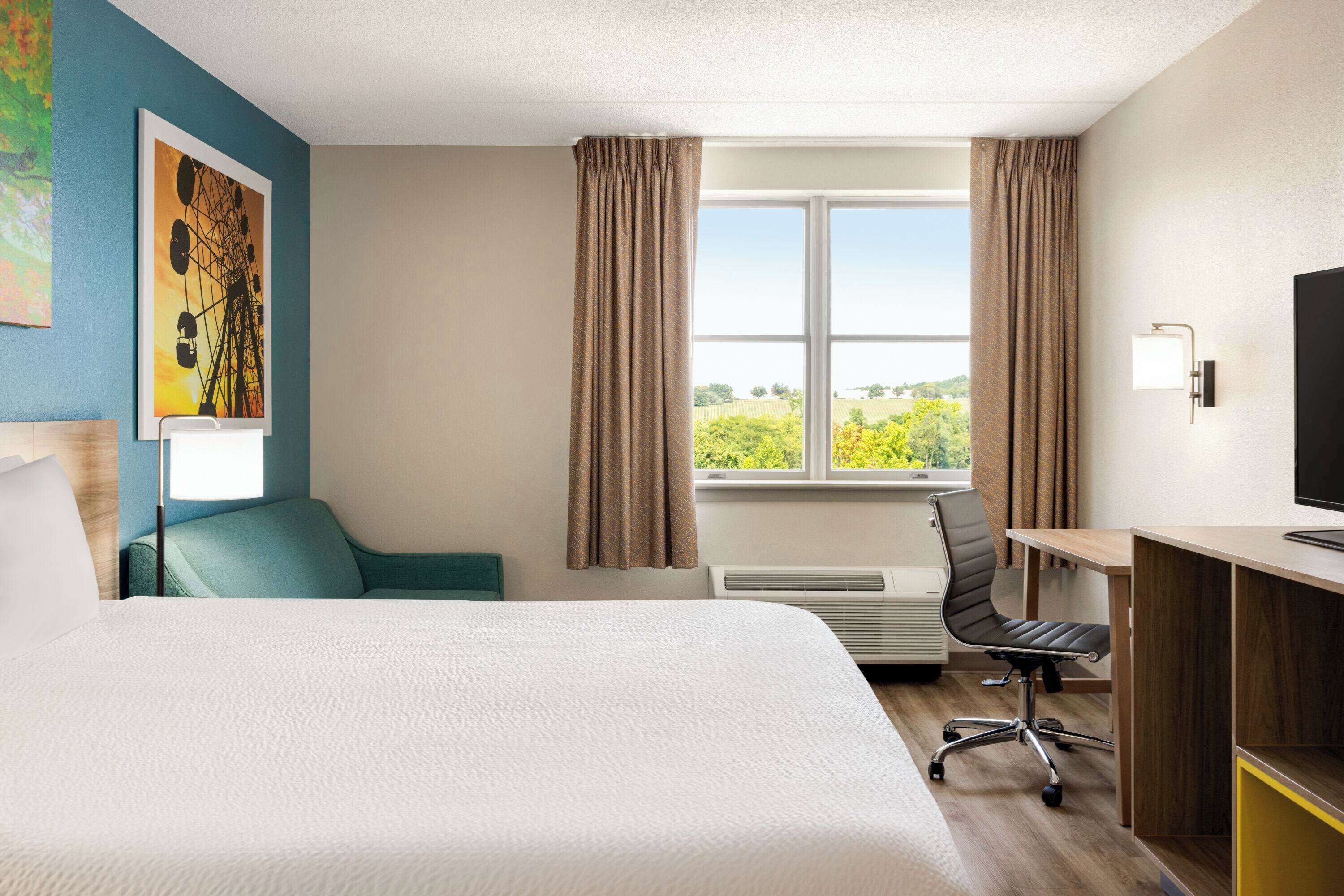 Una habitación de hotel con una cama, un escritorio con una silla, una ventana con cortinas y un cuadro en la pared.