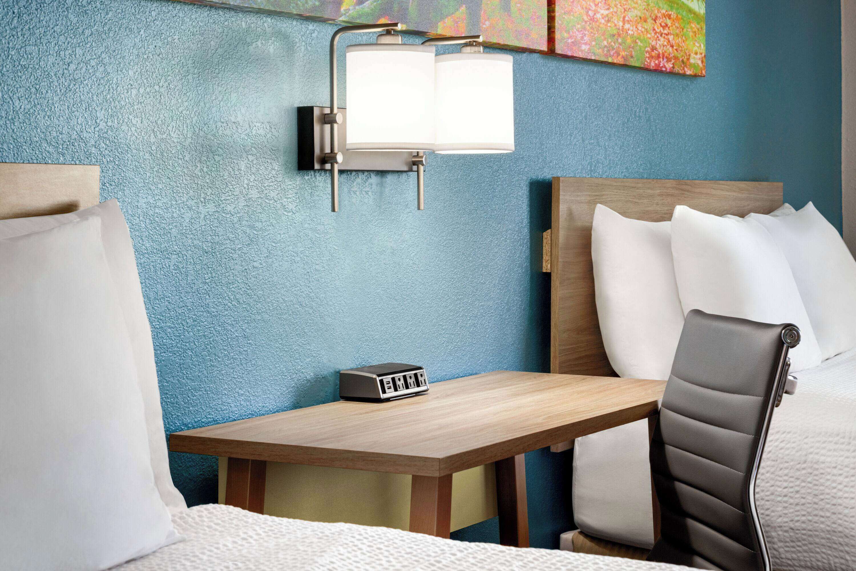 A hotel room with a bed, a desk with a clock, and a wall-mounted light fixture.