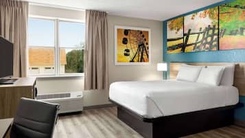 Ein Hotelzimmer mit Bett, Schreibtisch, Stuhl, einem Fenster mit Vorhängen, einer Klimaanlage und Bildern an der Wand.