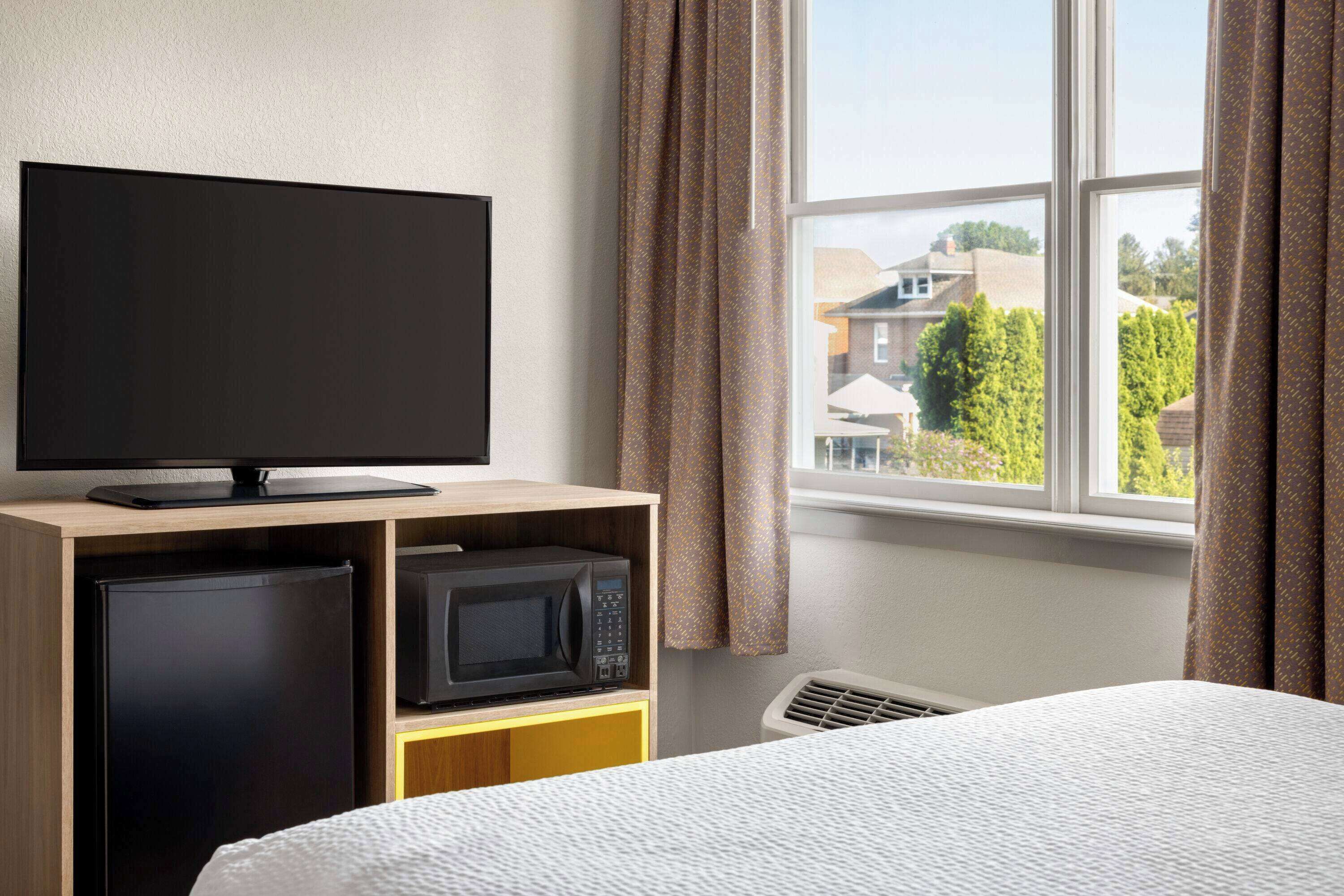 Habitación de hotel con cama, televisor en un estante de madera, microondas y ventana con cortinas.