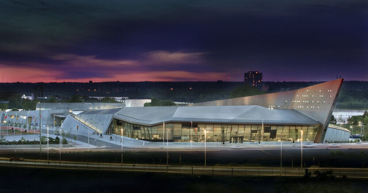 A modern building with a unique angular roof design, surrounded by a landscaped area with pathways and a body of water in the background.