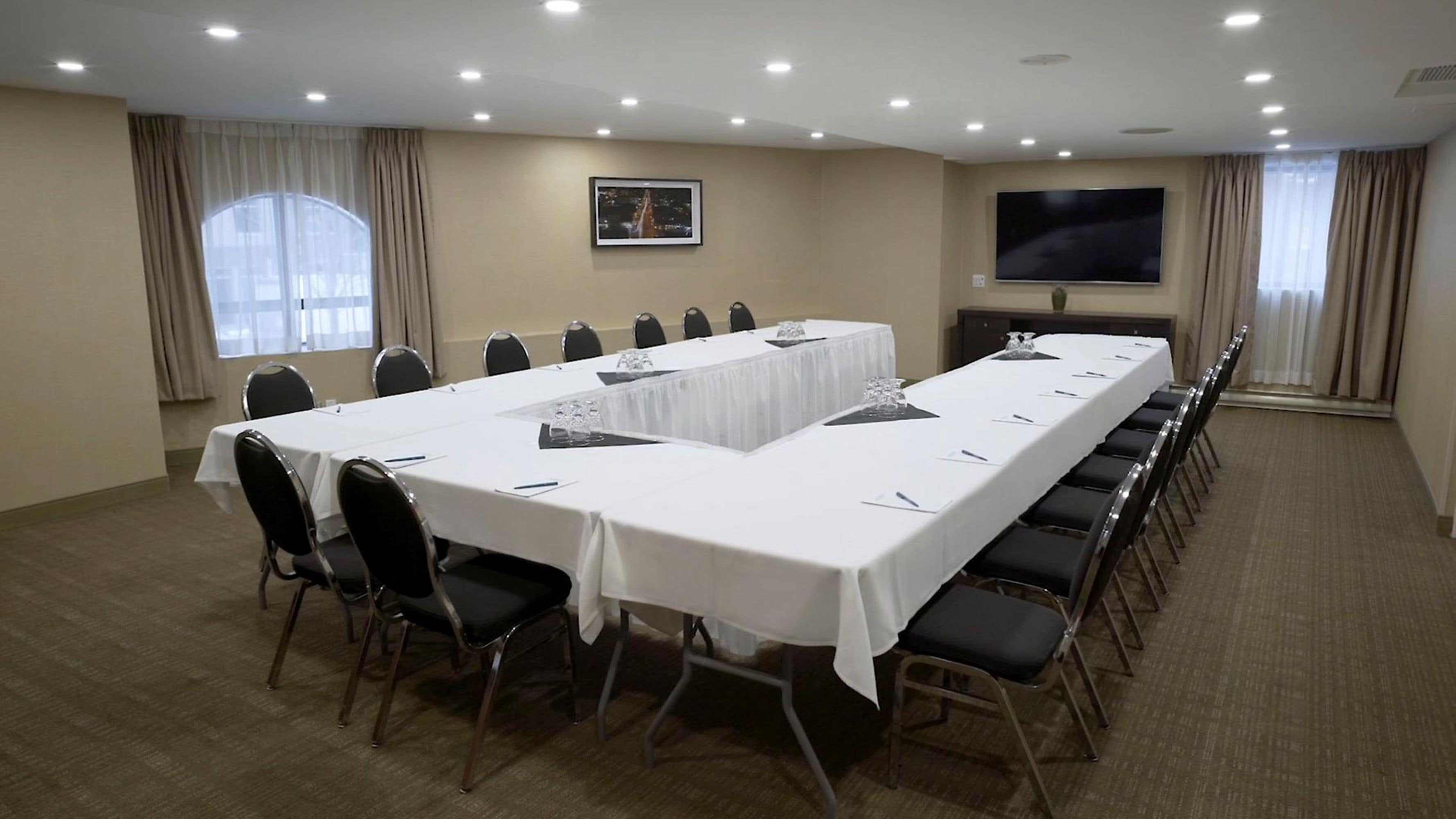 Un salón de conferencias con una larga mesa, sillas, un televisor de pantalla plana y ventanas con cortinas.