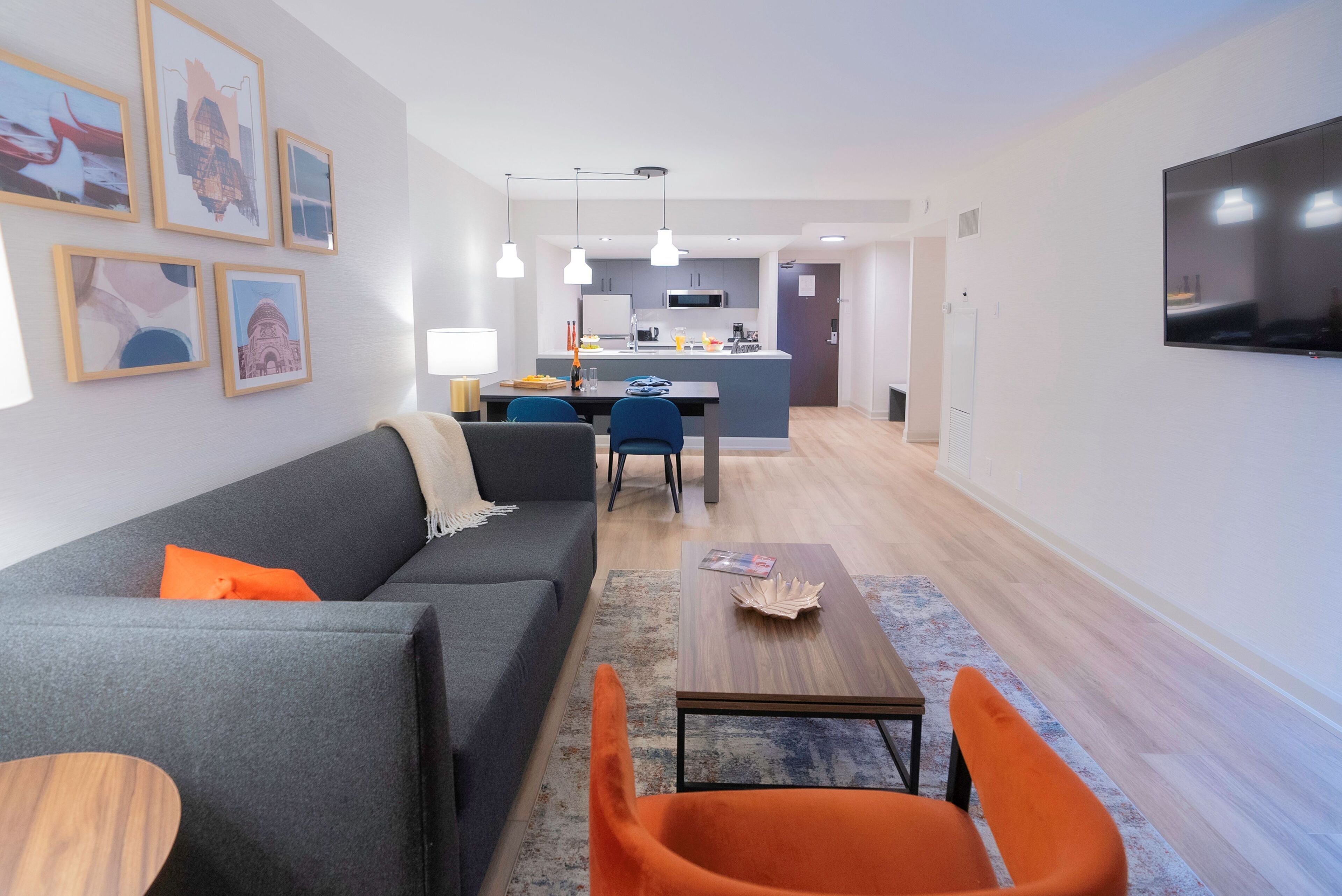 A modern living room with a grey sofa, orange armchairs, and a wooden coffee table. A kitchen is visible in the background with white cabinets and a flat-screen TV mounted on the wall.
