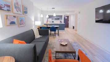 Una sala de estar moderna con un sofá gris, sillones naranjas y una mesa de centro de madera. Se ve una cocina al fondo, con armarios blancos y un televisor de pantalla plana montado en la pared.