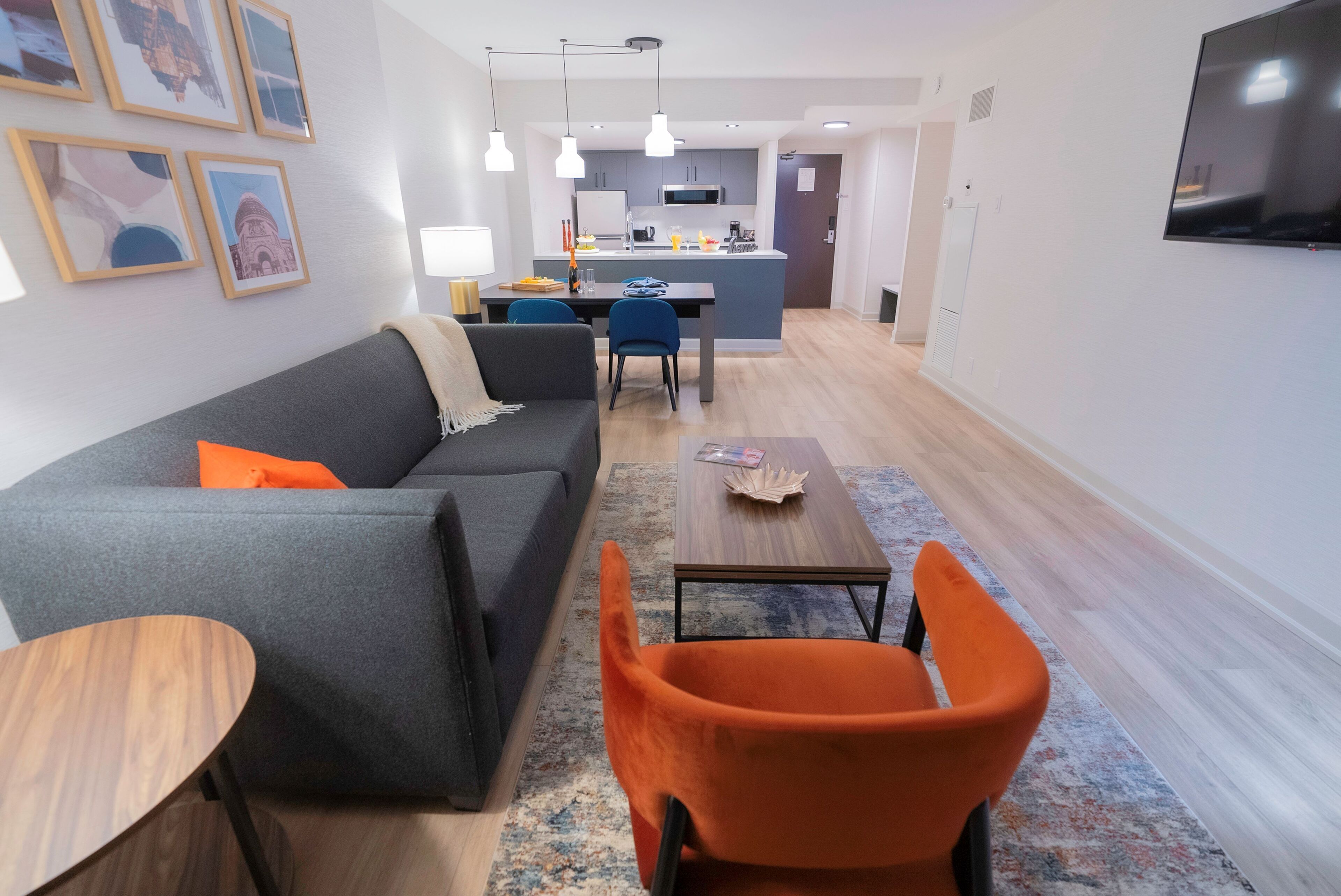 A modern living room with a grey sofa, orange armchair, and a small wooden table. There is a kitchen area in the background with a microwave and cabinets.