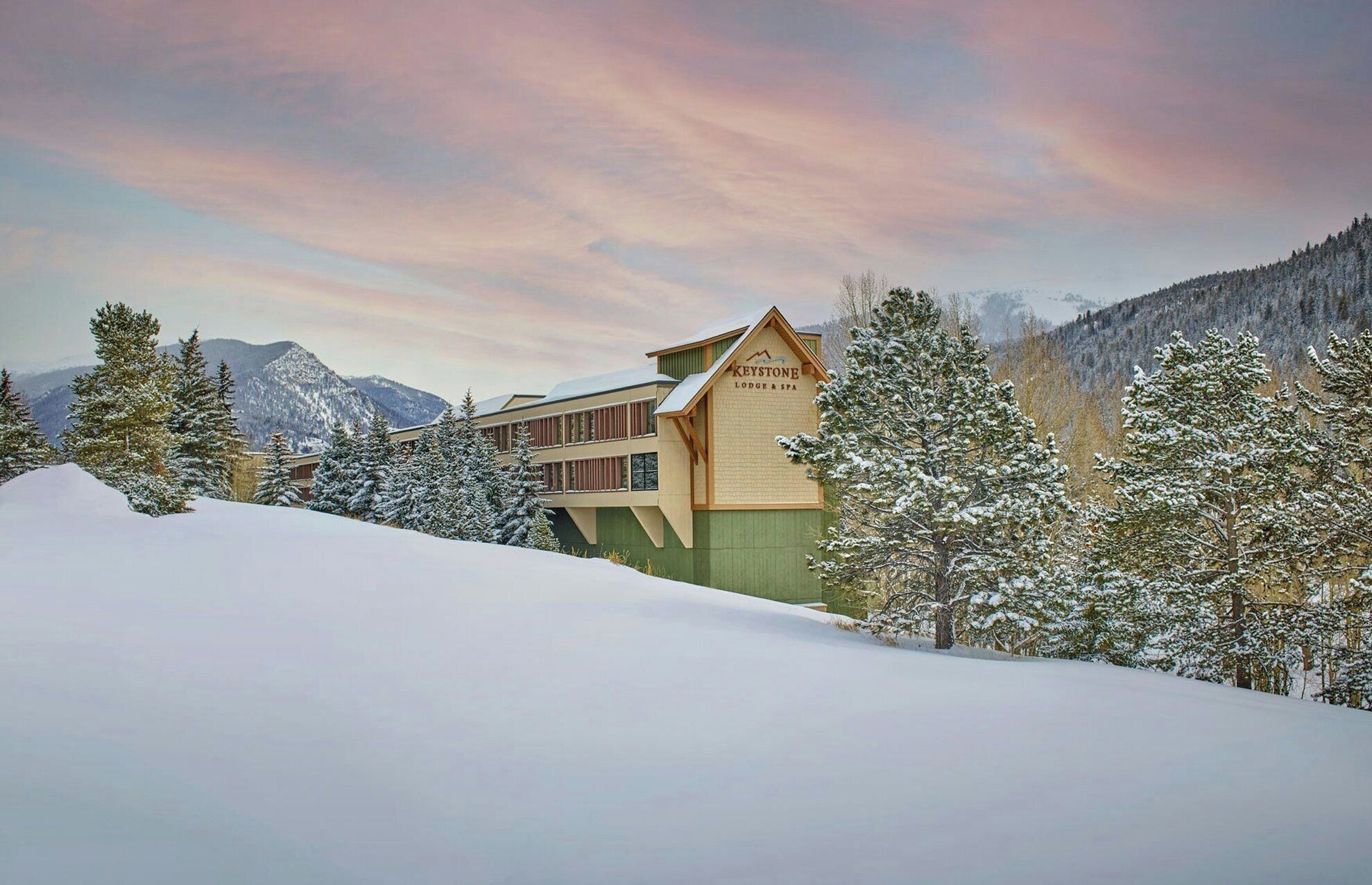 A lodge with a sign reading "Keystone Lodge & Spa" amidst snow-covered trees and mountains.
