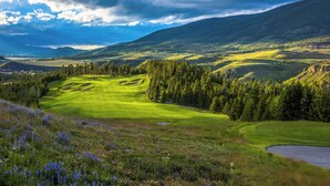 A golf course with rolling hills, dense forests, and a cloudy sky.