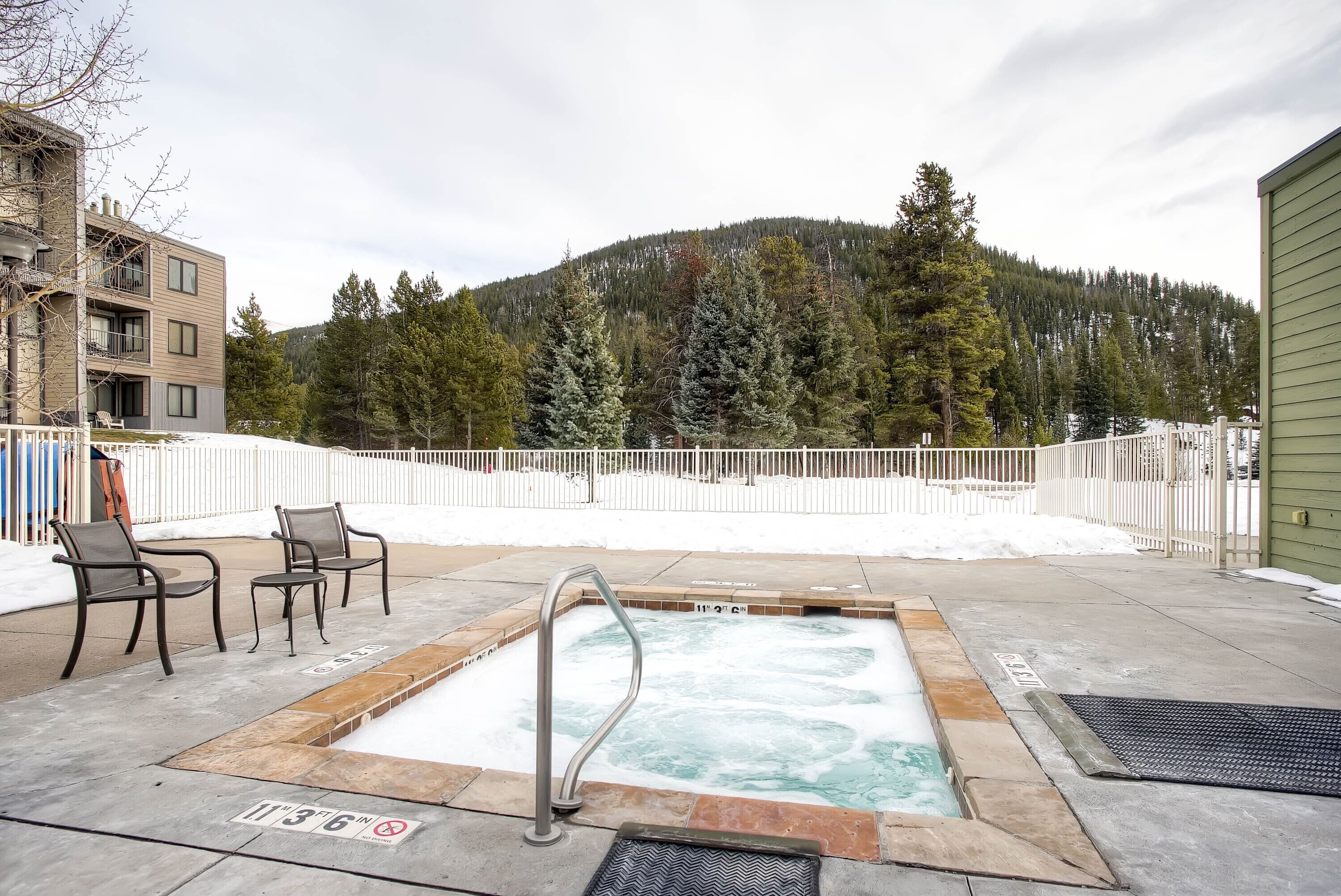 A hot tub area with a view of a forested mountain, outdoor seating, and a building with balconies.