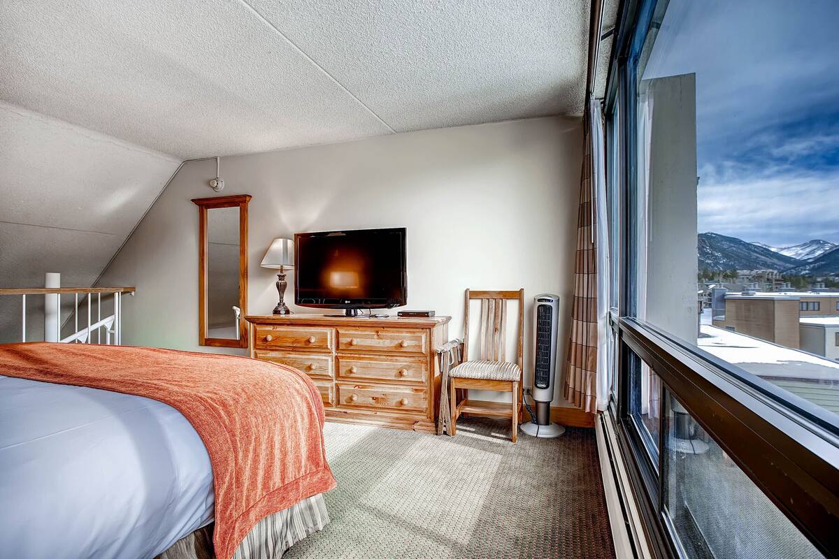 A hotel room with a bed, a wooden dresser, a television, a chair, and a window with a mountain view.