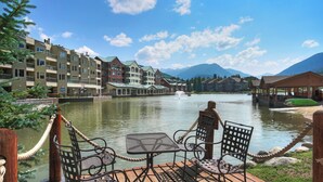 A lakeside deck with outdoor seating, overlooking a calm lake and a row of multi-story buildings with balconies.