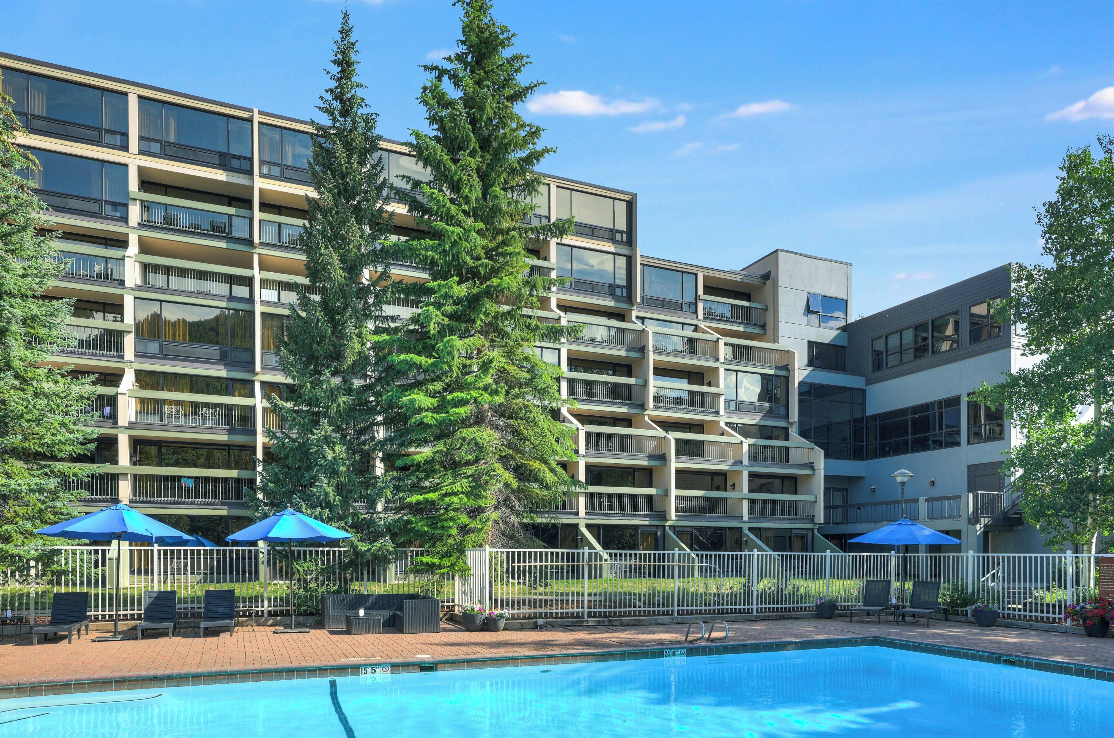Um edifício moderno de vários andares com piscina, área de estar ao ar livre e uma grande árvore na frente.