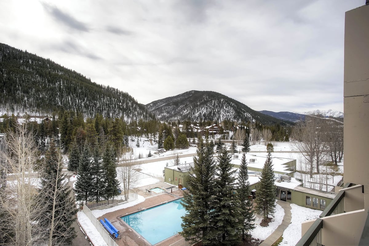 A view of a residential area with a swimming pool, surrounded by snow-covered ground and trees, with mountains in the background.
