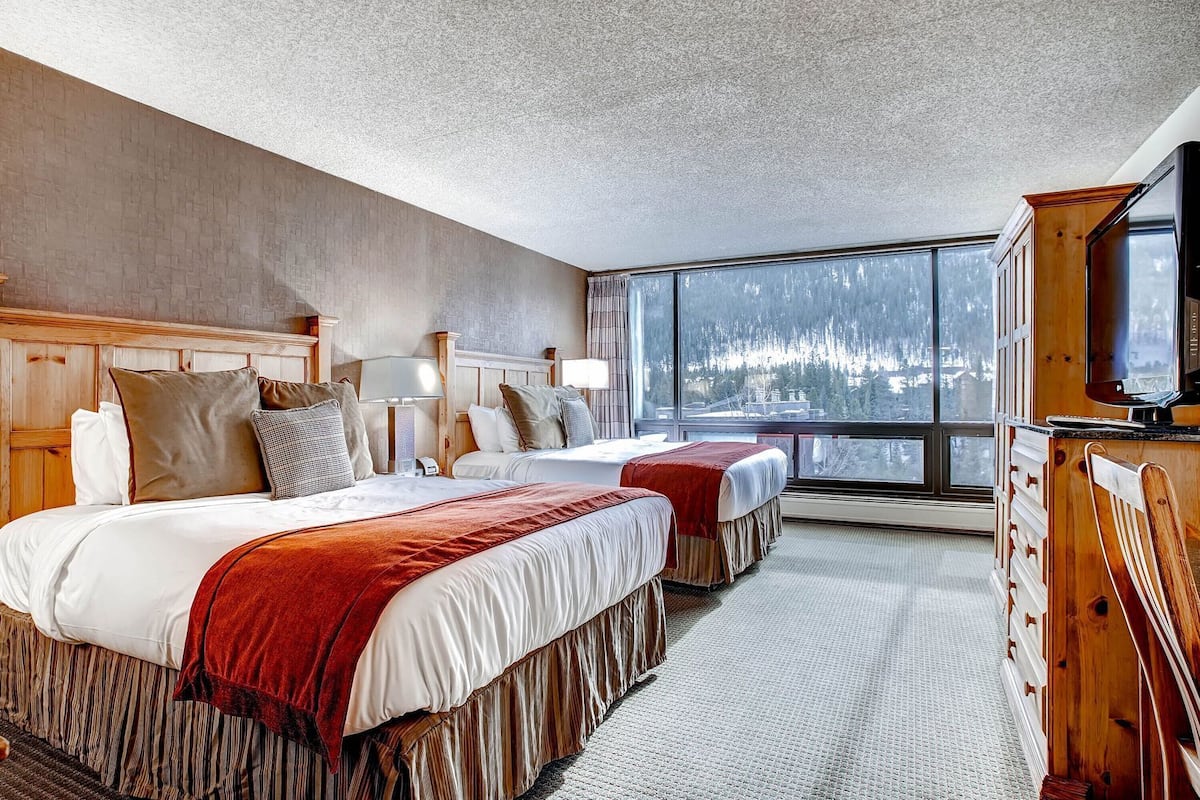 A hotel room with two beds, a large window with a view of a snowy landscape, a wooden dresser, and a television.