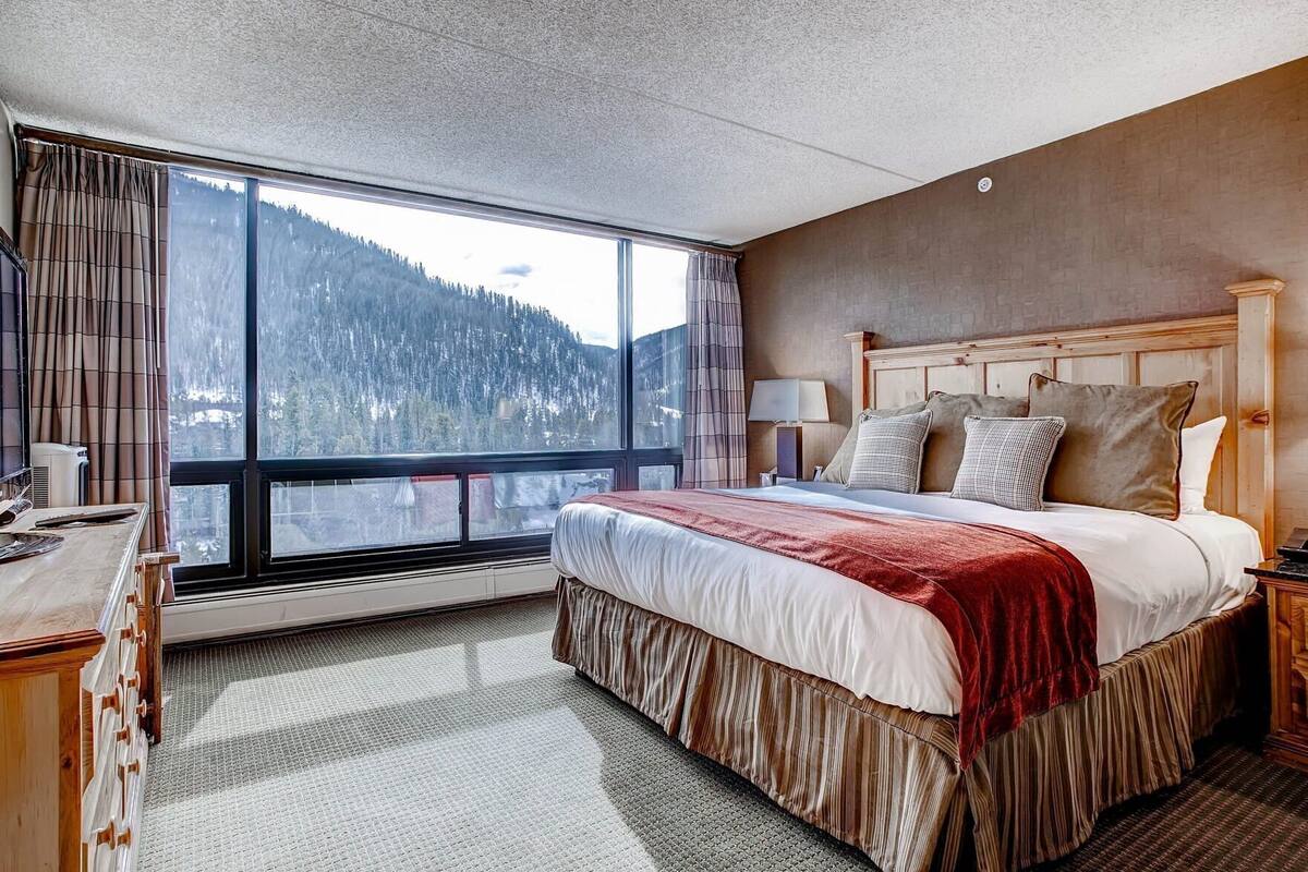 A hotel room with a large window, a bed with a red blanket, a wooden headboard, and a desk with a lamp.