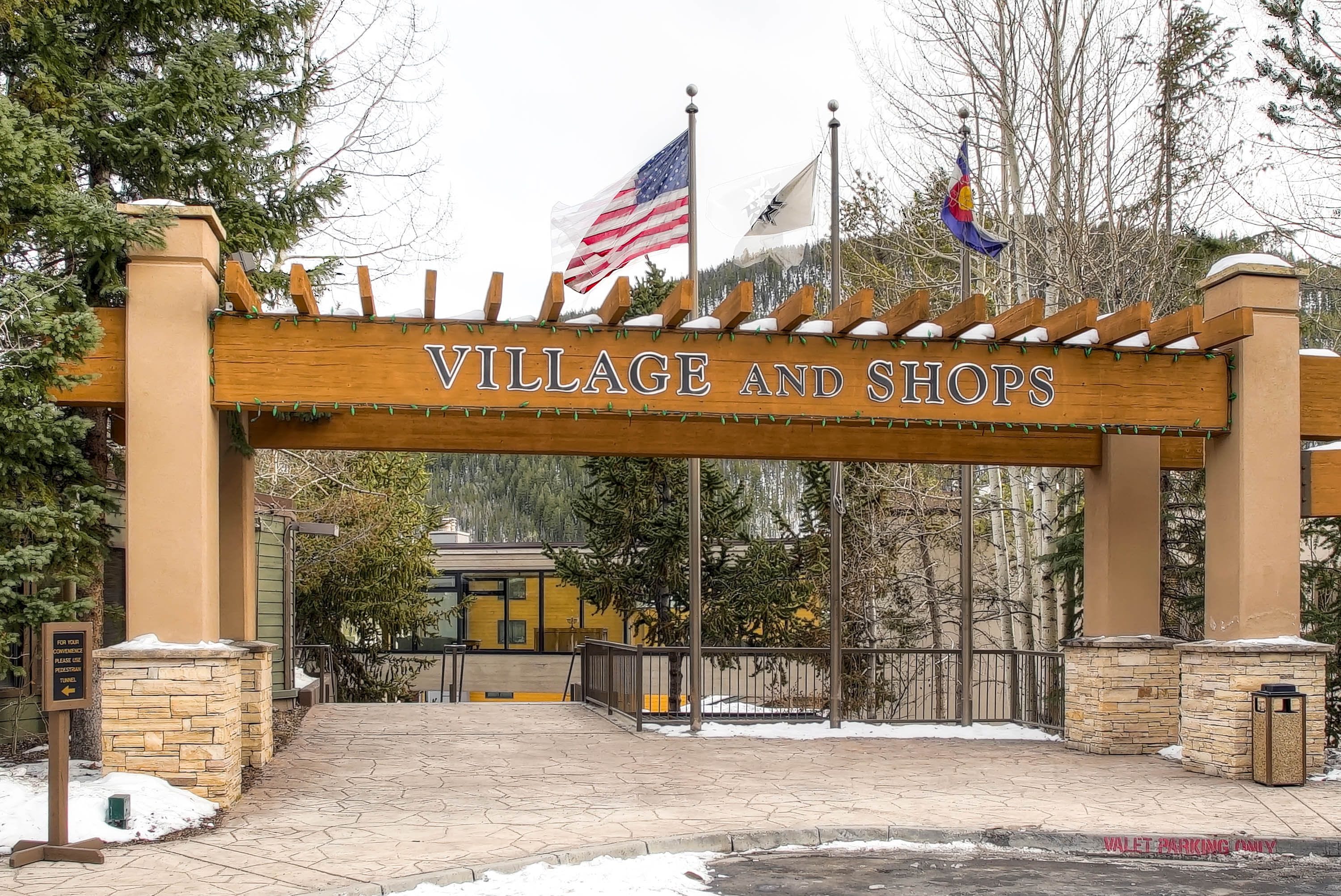 A wooden archway with "VILLAGE AND SHOPS" written on it, flanked by American and another flag, leading to a building with a yellow facade.