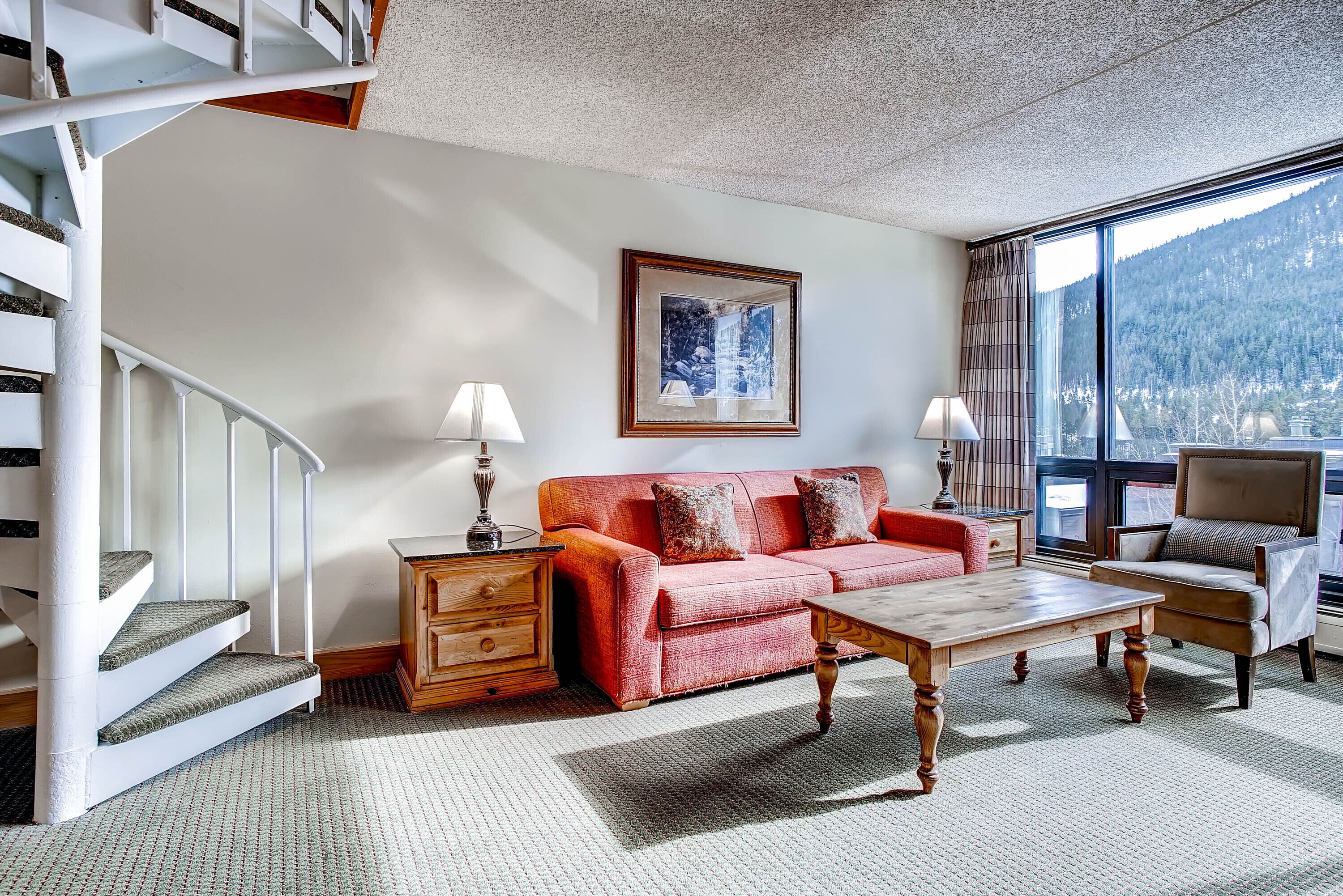 A living room with a staircase, a sofa, a coffee table, and a view of a mountain.