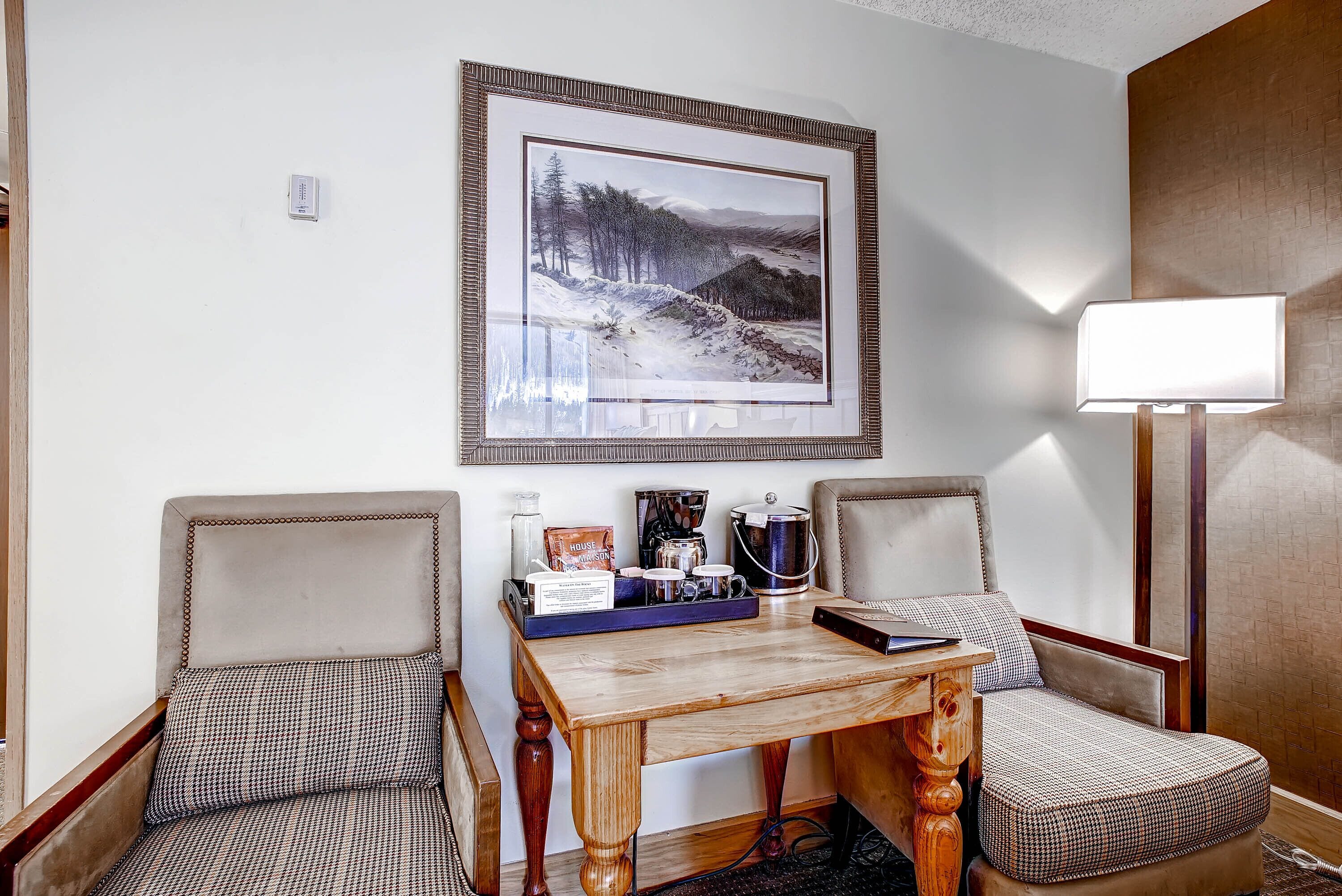 Habitación de hotel con dos sillones, una mesa de madera, un cuadro enmarco de paisaje, una zona de café y una lámpara de noche.