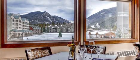 Un coin repas avec une table dressée pour deux personnes, de grandes fenêtres offrant une vue sur les montagnes et un vase décoratif posé sur la table.