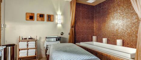 A hotel room with a bed, a vanity with a mirror, a shelf with towels, a trash bin, and a wall with a mosaic pattern.