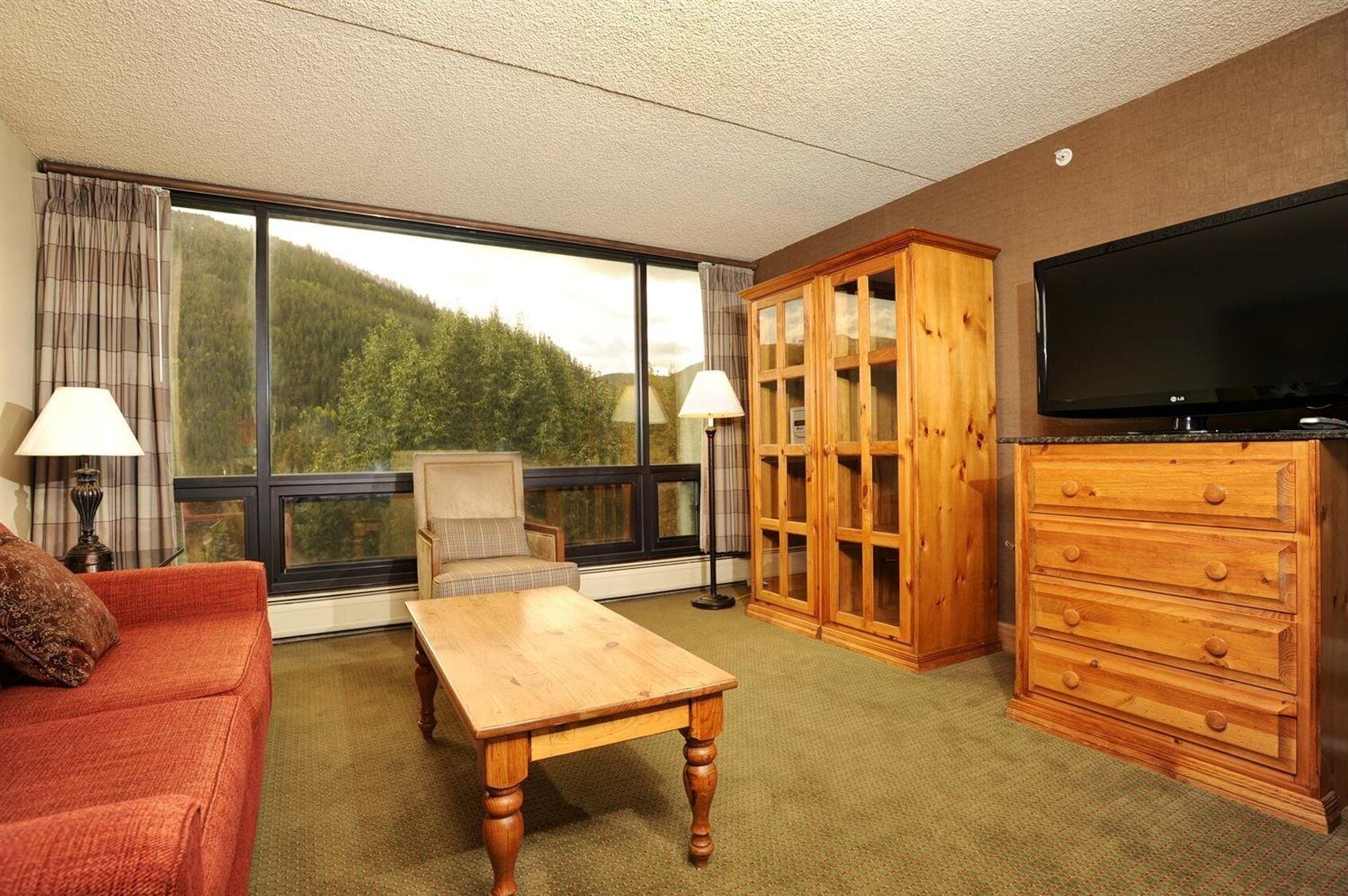 A hotel room with a large window, a wooden cabinet, a television, a sofa, a chair, and a table.