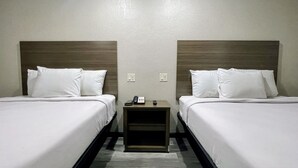 Ein Hotelzimmer mit zwei Einzelbetten, jeweils mit weißer Bettwäsche und Kissen. An den Nachttischen stehen je ein Telefon und eine Fernbedienung. Die Wände sind in einer hellen Farbe gestrichen und es sind zwei Steckdosen an der Wand sichtbar.