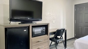 Ein kleines Zimmer mit einem Flachbildfernseher auf einer Holzkommode, einer Mikrowelle, einem Sessel und einer Tür.