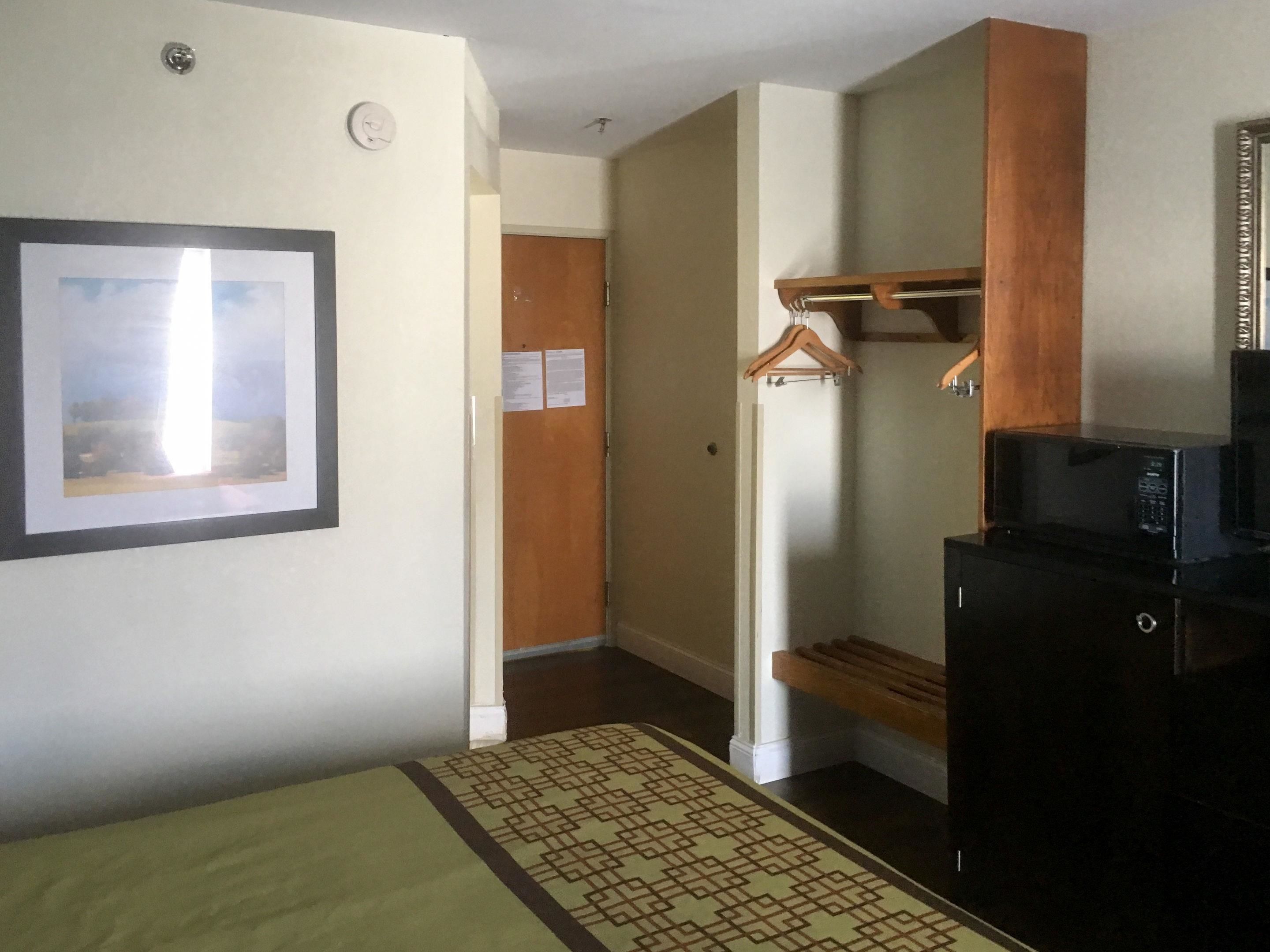 Habitación de hotel con cama, televisor montado en la pared, armario de madera, microondas y perchero.