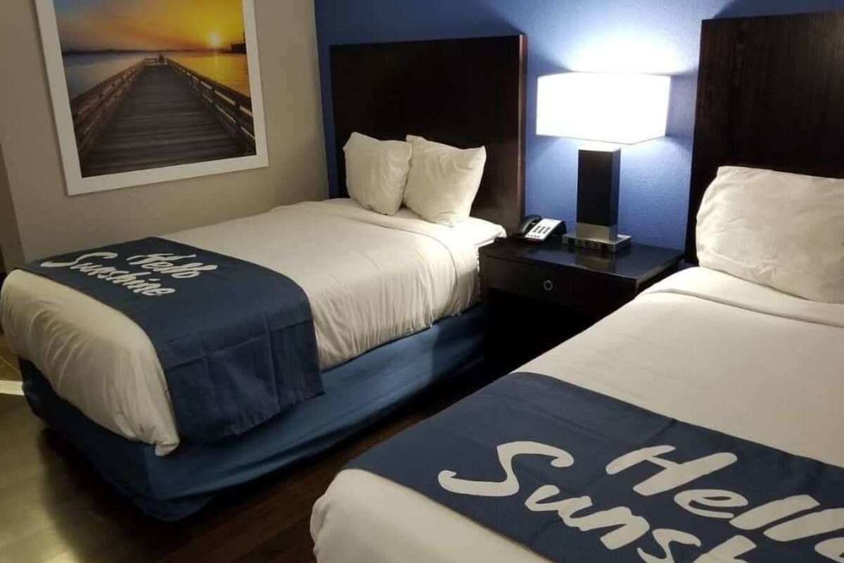 Two beds with white linens and blue headboards, a nightstand with a lamp, and a framed picture of a pier on the wall.