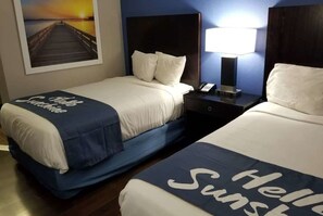 Two beds with white linens and blue headboards, a nightstand with a lamp, and a framed picture of a pier on the wall.
