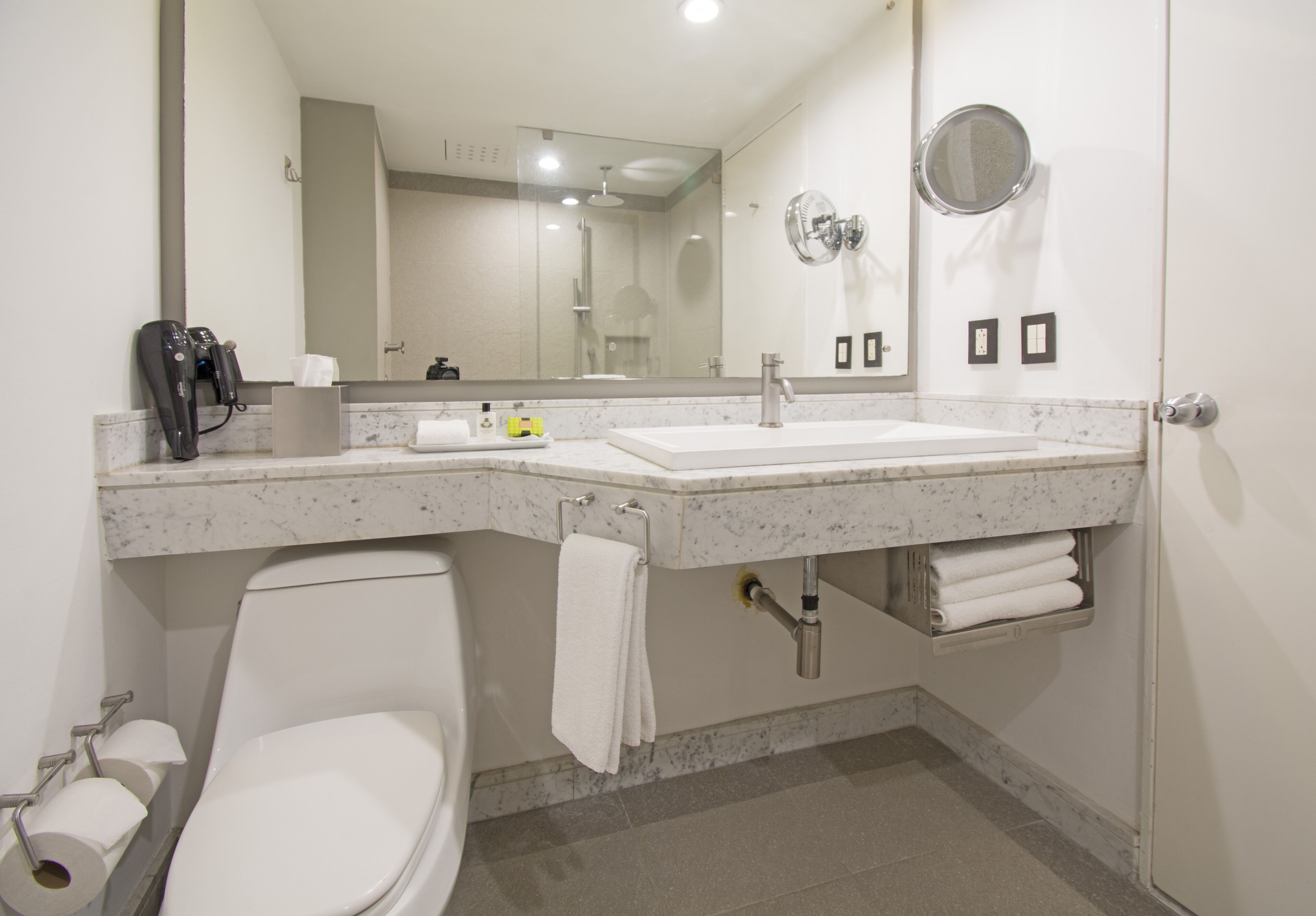 Un baño con inodoro blanco, lavamanos, espejo y toallero.