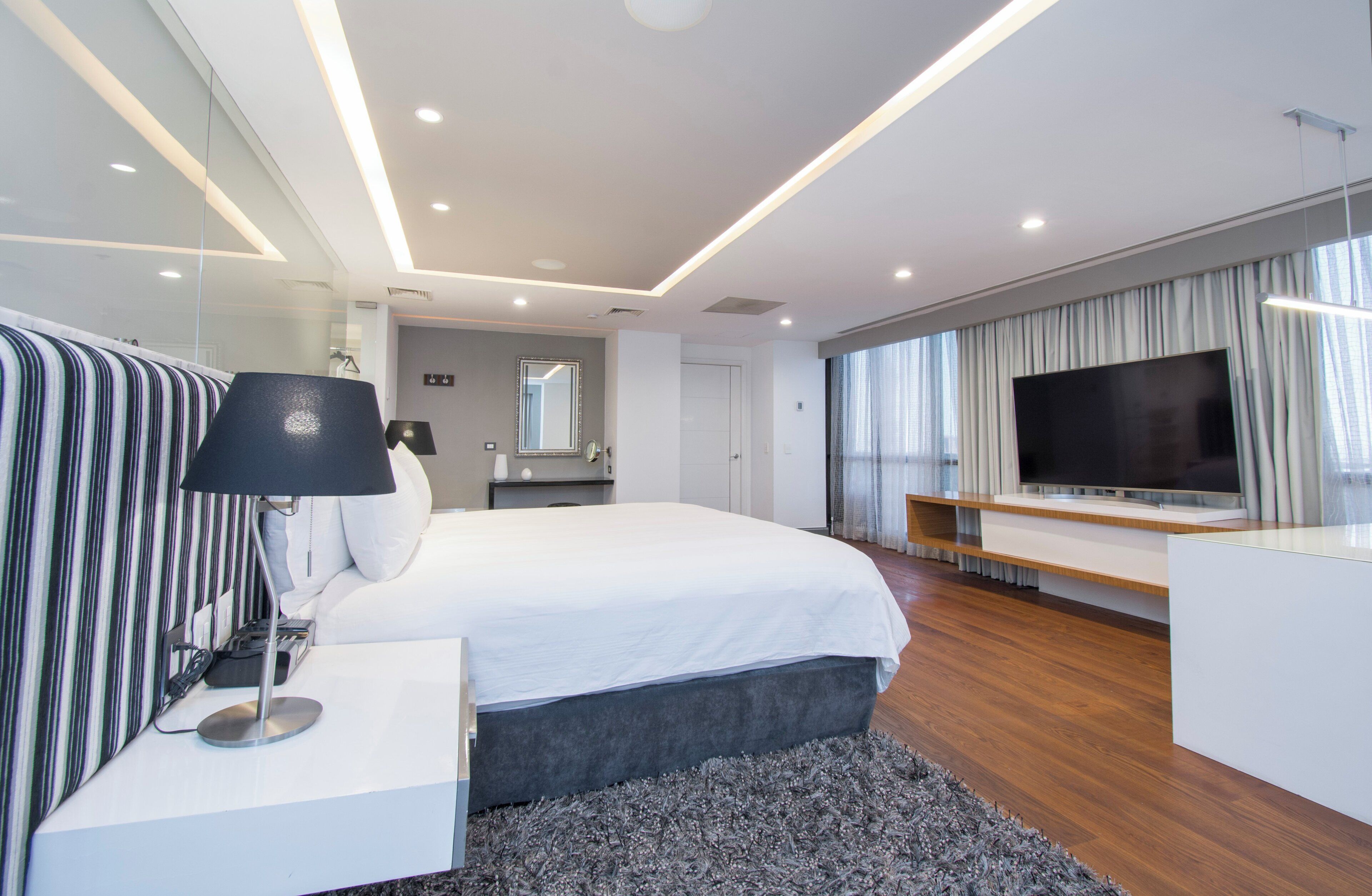 Una habitación de hotel moderna con una cama grande, un televisor de pantalla plana, una mesita de noche con lámpara y un espejo amplio.