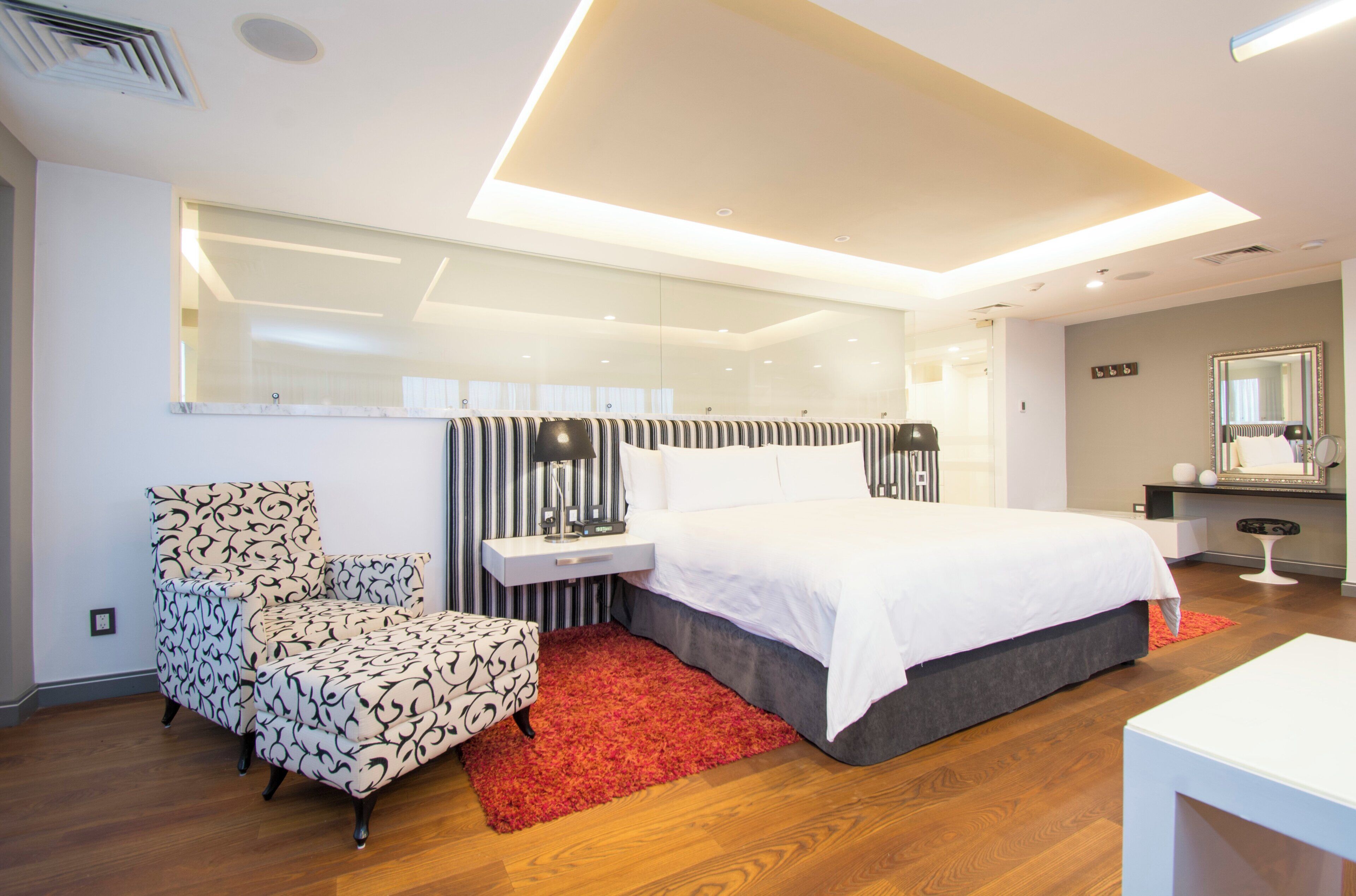 Habitación de hotel moderna con una cama grande, un rincón de descanso con un sillón estampado, una mesita de noche con lámpara y un espejo en la pared.